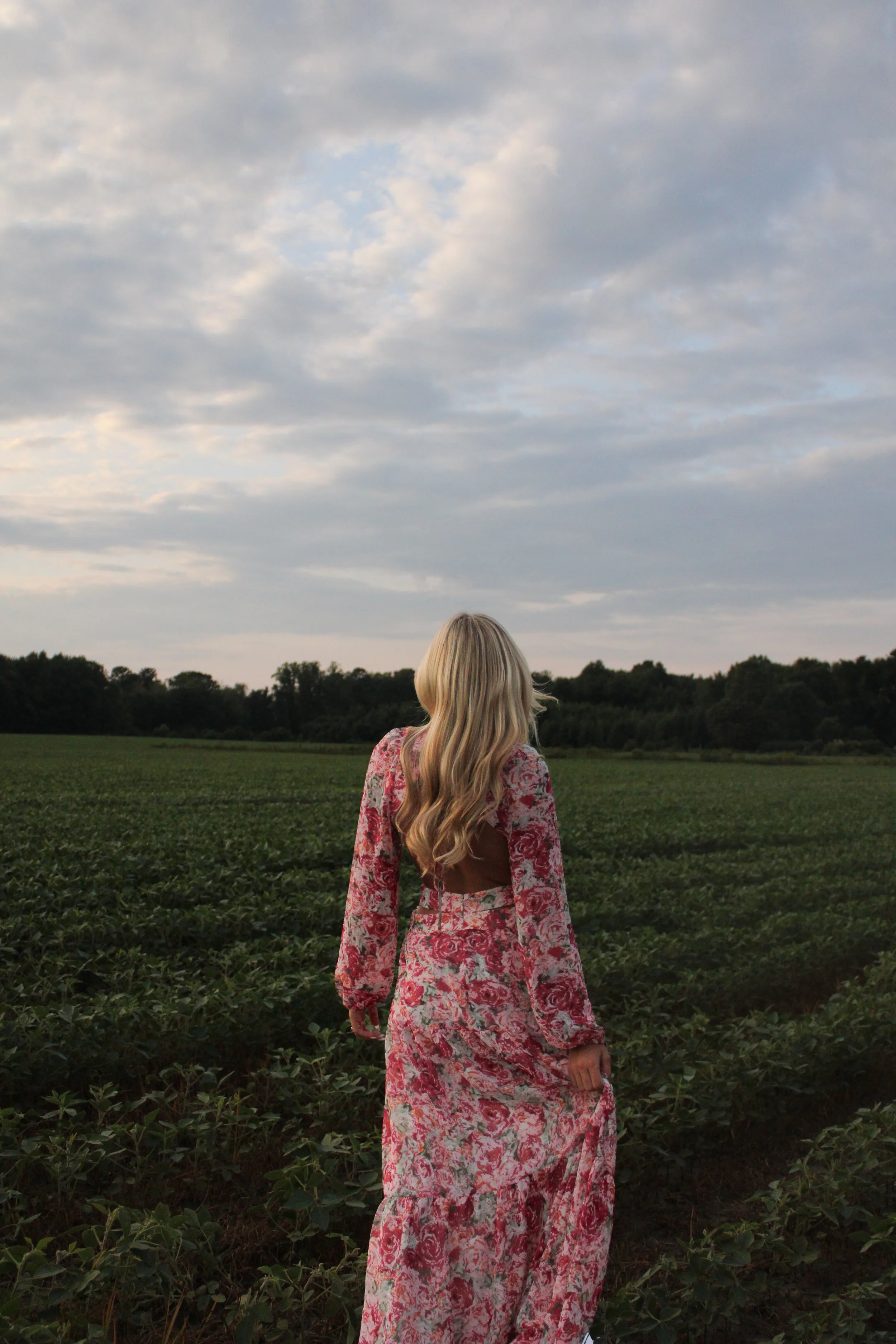 A woman with long blonde hair wearing a pink floral dress standing in a green field, facing away from the camera, under a cloudy sky.
