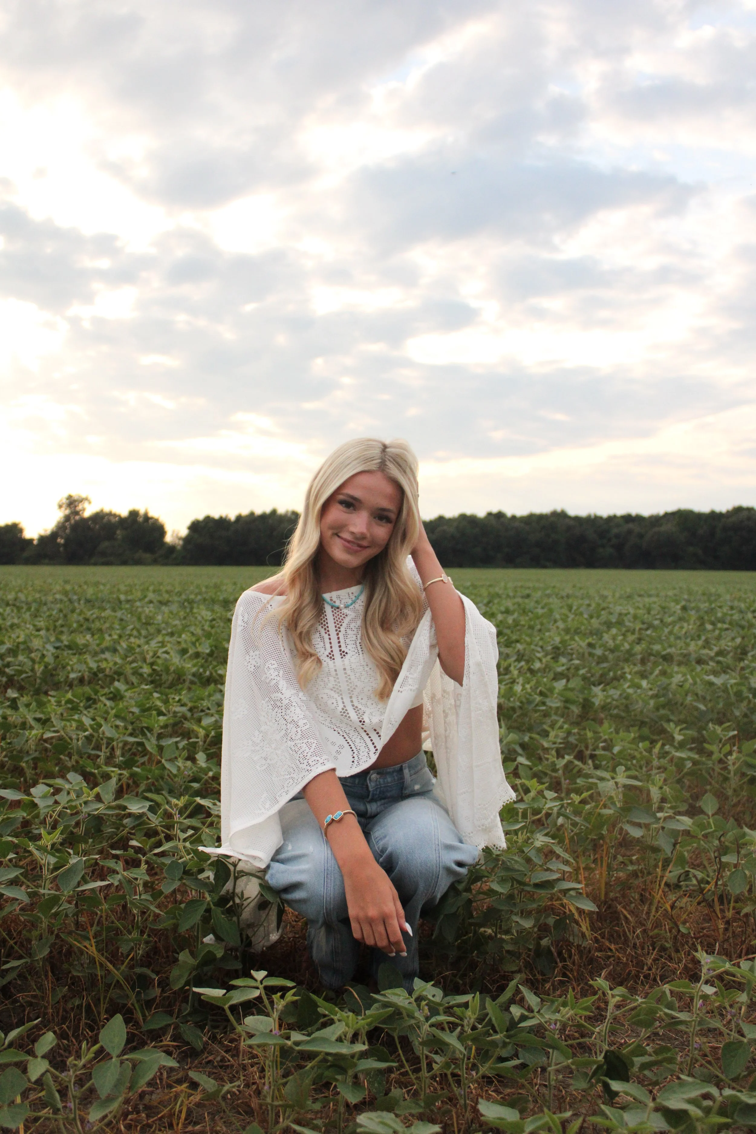 A young woman with long blonde hair sitting in a green field of crops under a partly cloudy sky. She is smiling, wearing a white crochet top, blue jeans, and has bracelets on her left wrist.