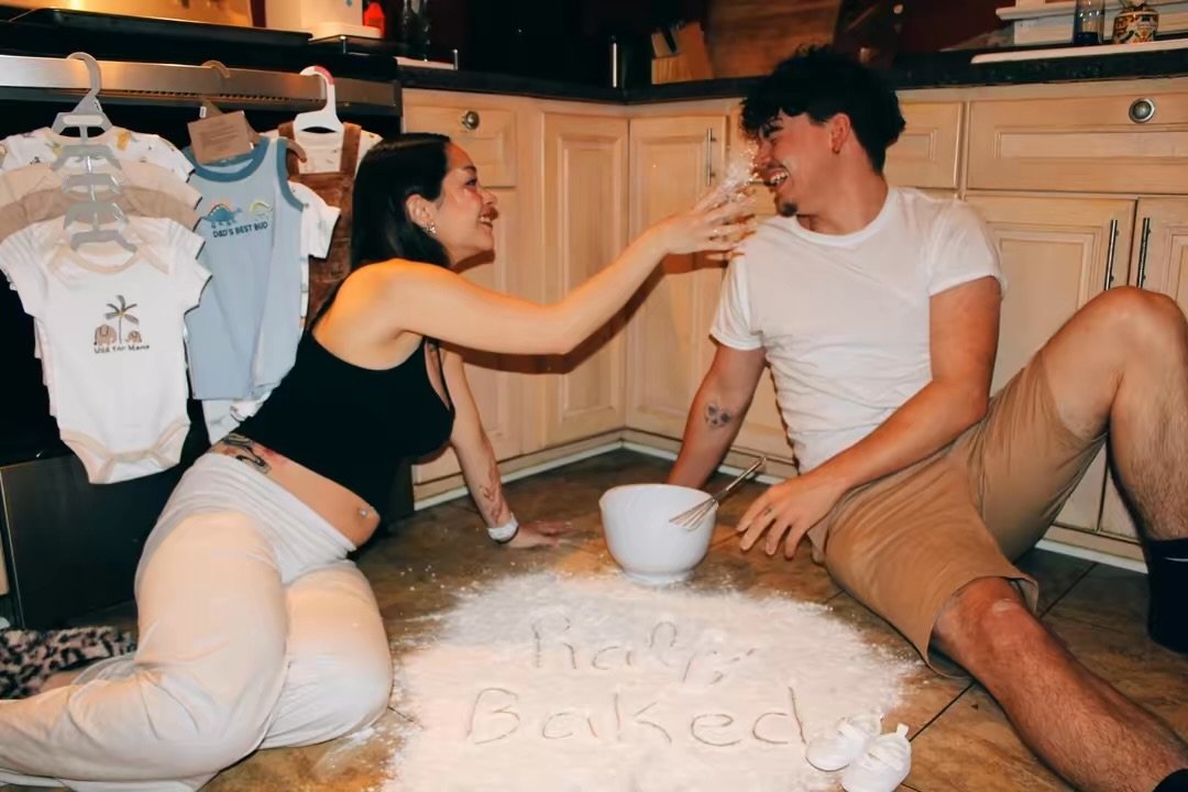 A woman teasing a man with a glass of water while sitting on a kitchen floor, with 'I'm Baked' written in flour on the floor, and baby clothes hanging on a rack in the background.
