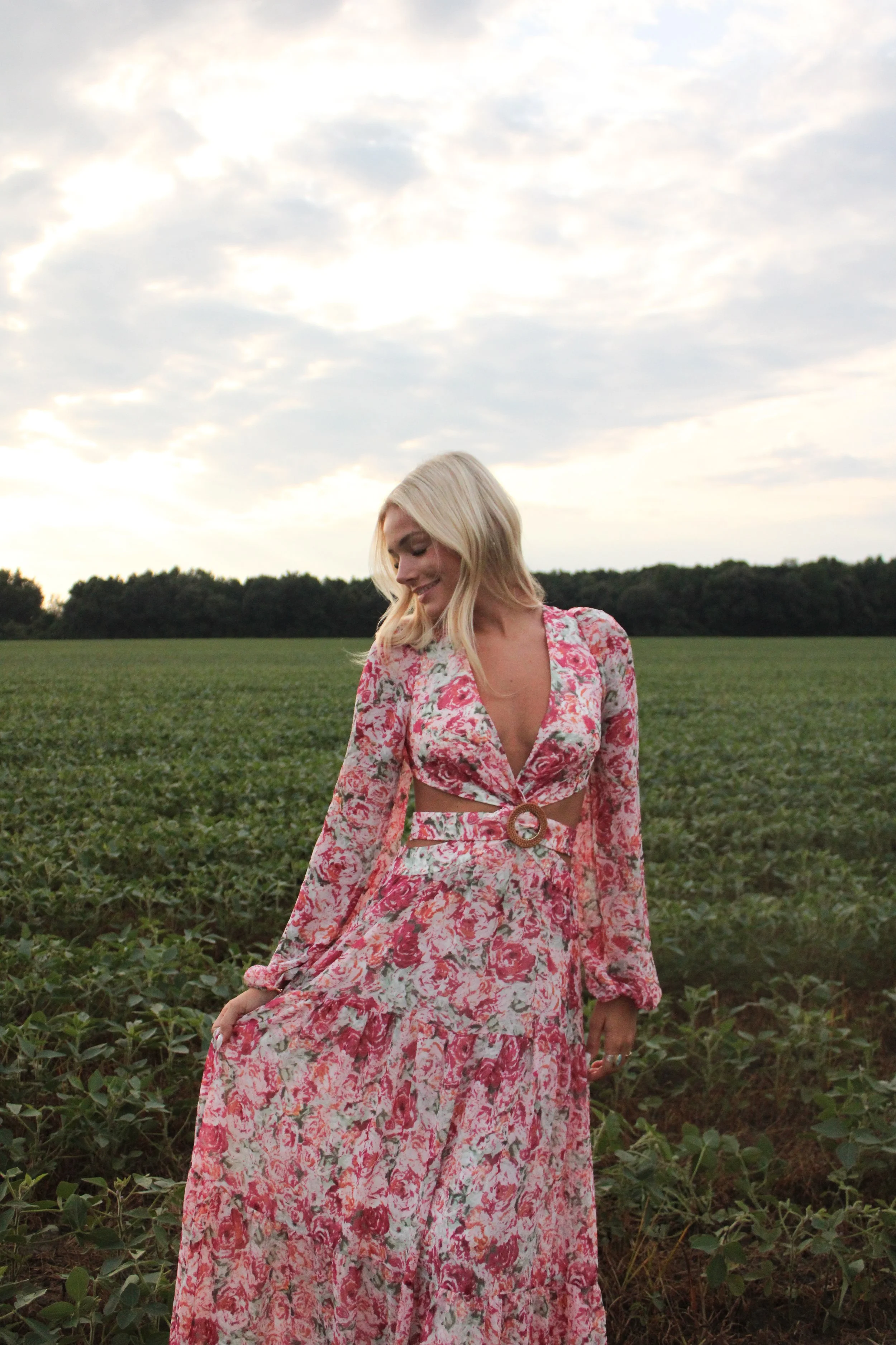A young woman with blonde hair wearing a pink floral dress standing in a green field with trees in the background and a cloudy sky above.
