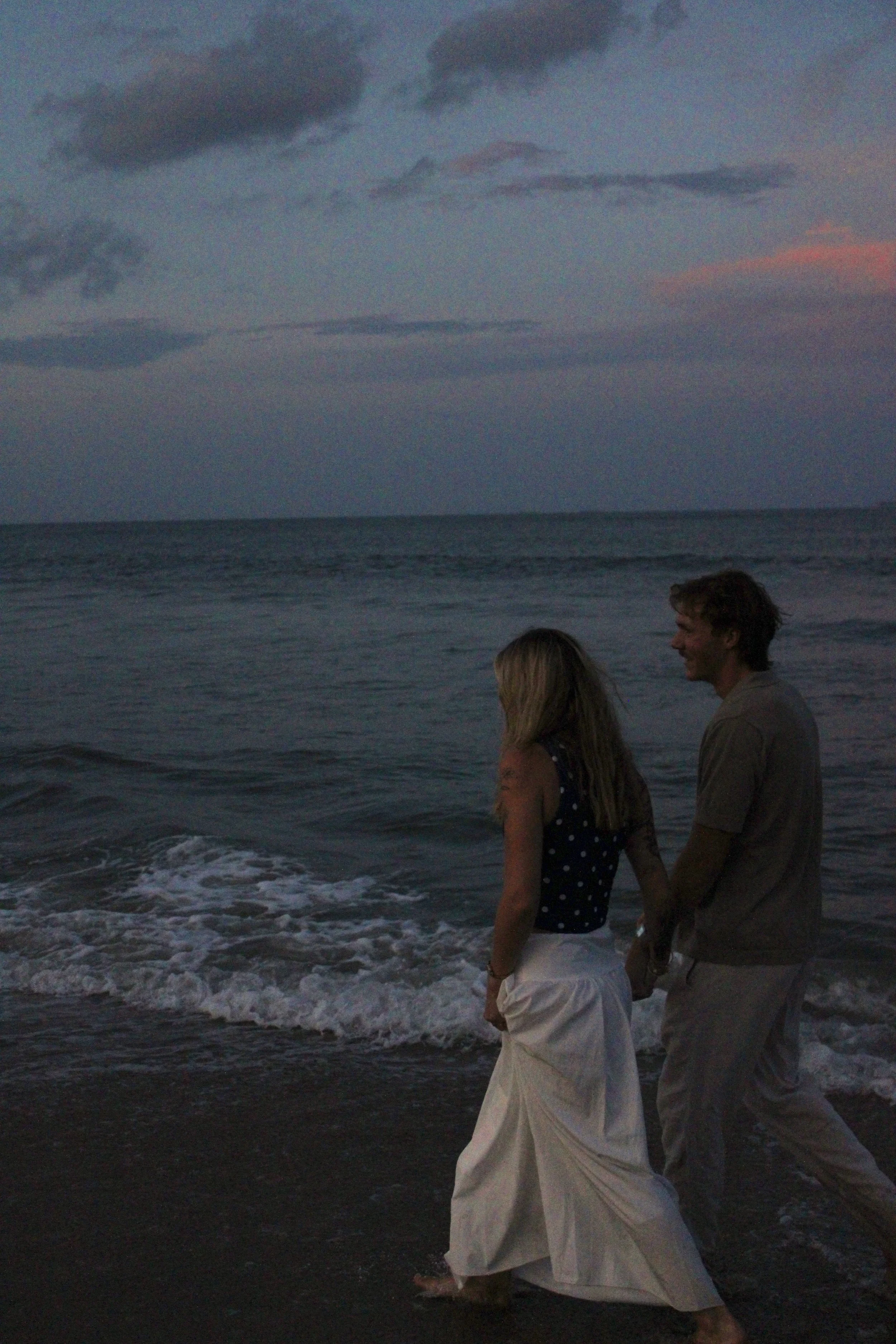A couple walking hand in hand along the shoreline at sunset or dusk on a beach, with waves gently crashing at their feet and cloudy sky above.