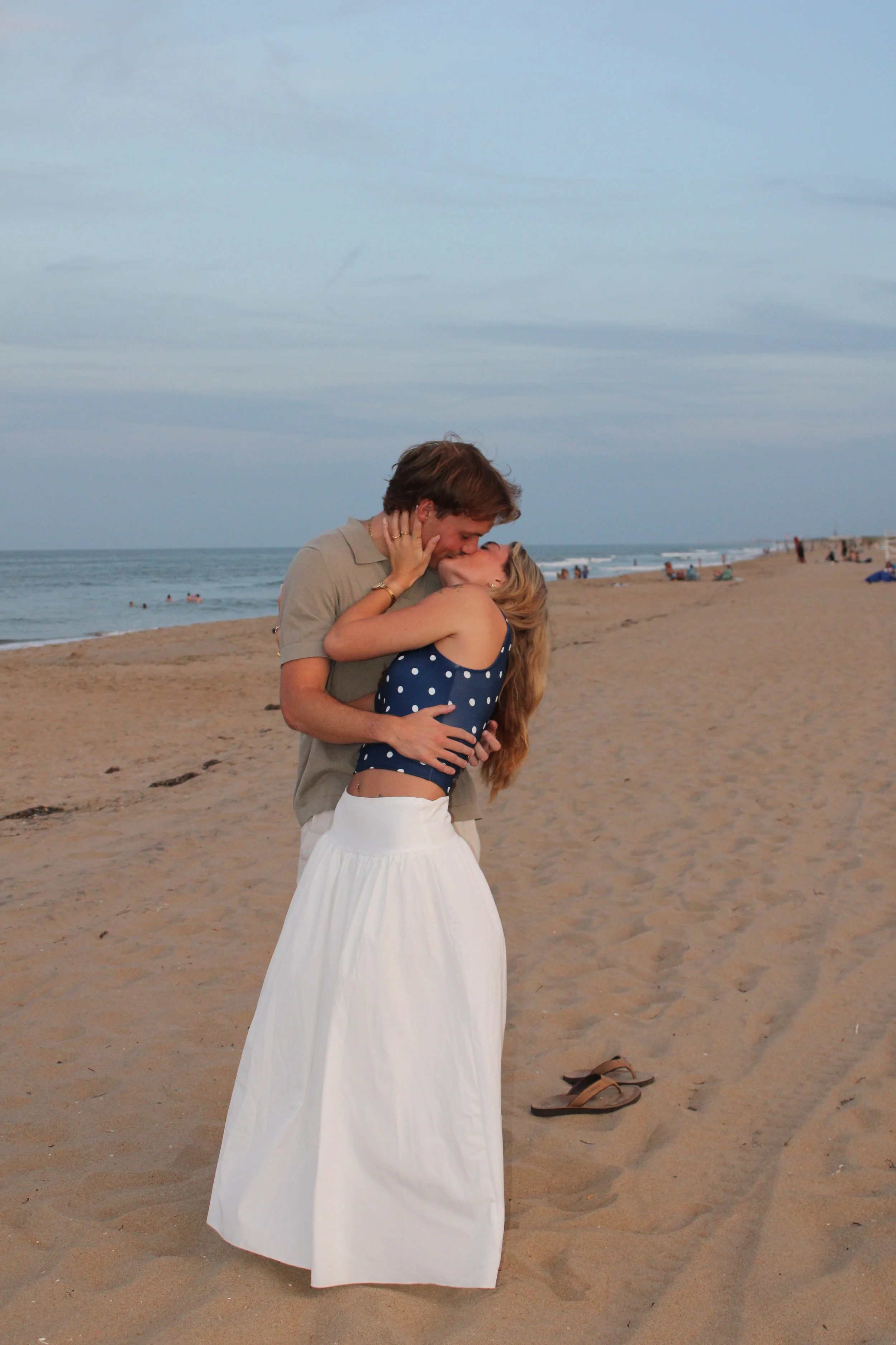 A couple sharing a kiss on the beach, with the man holding the woman and both smiling. The woman is wearing a blue polka dot top and a white skirt, and the man is dressed in a beige shirt. There are sandals on the sand near them and a cloudy sky over
