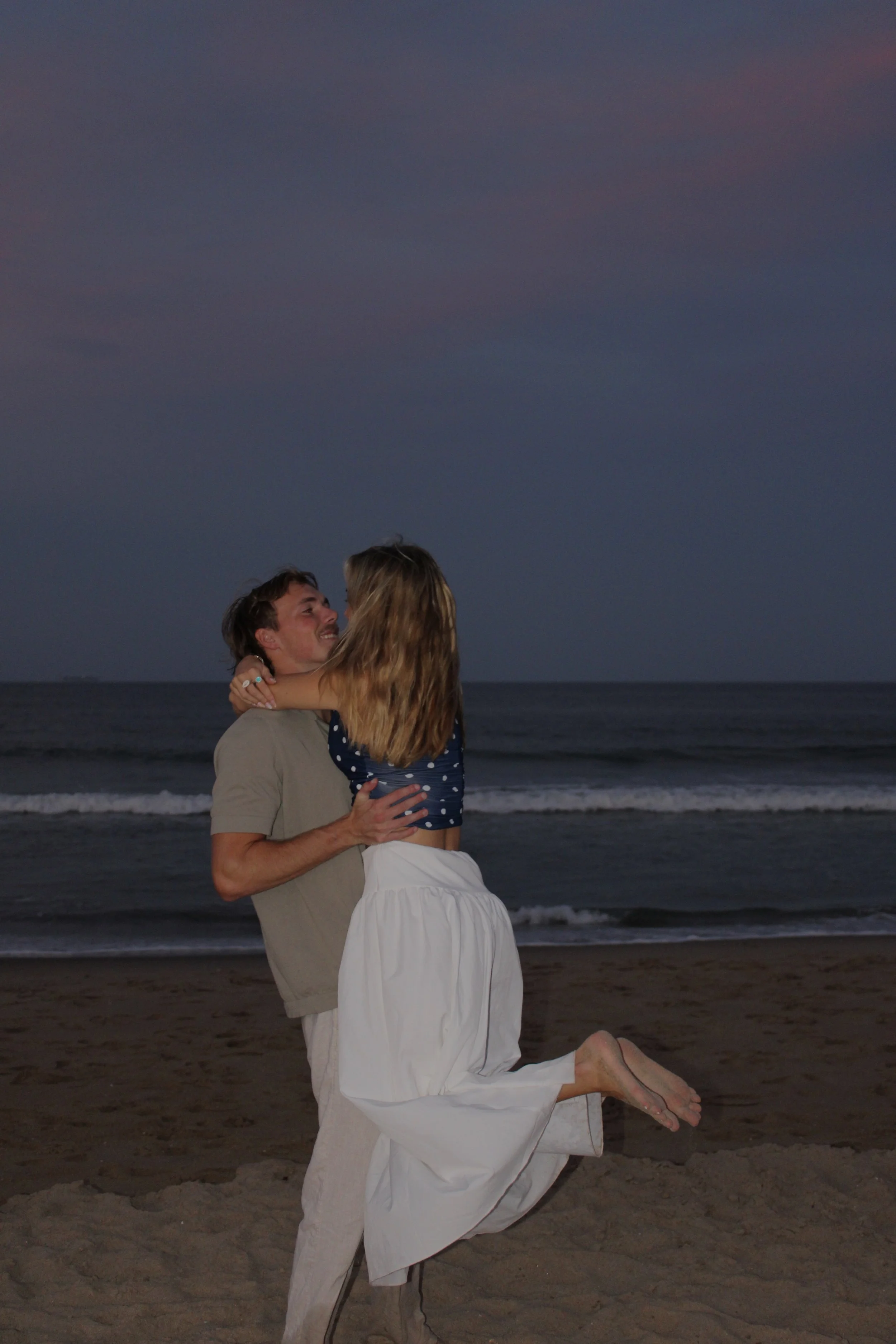 A couple at the beach during sunset, with the man kneeling and holding the woman, both smiling and looking into each other's eyes.