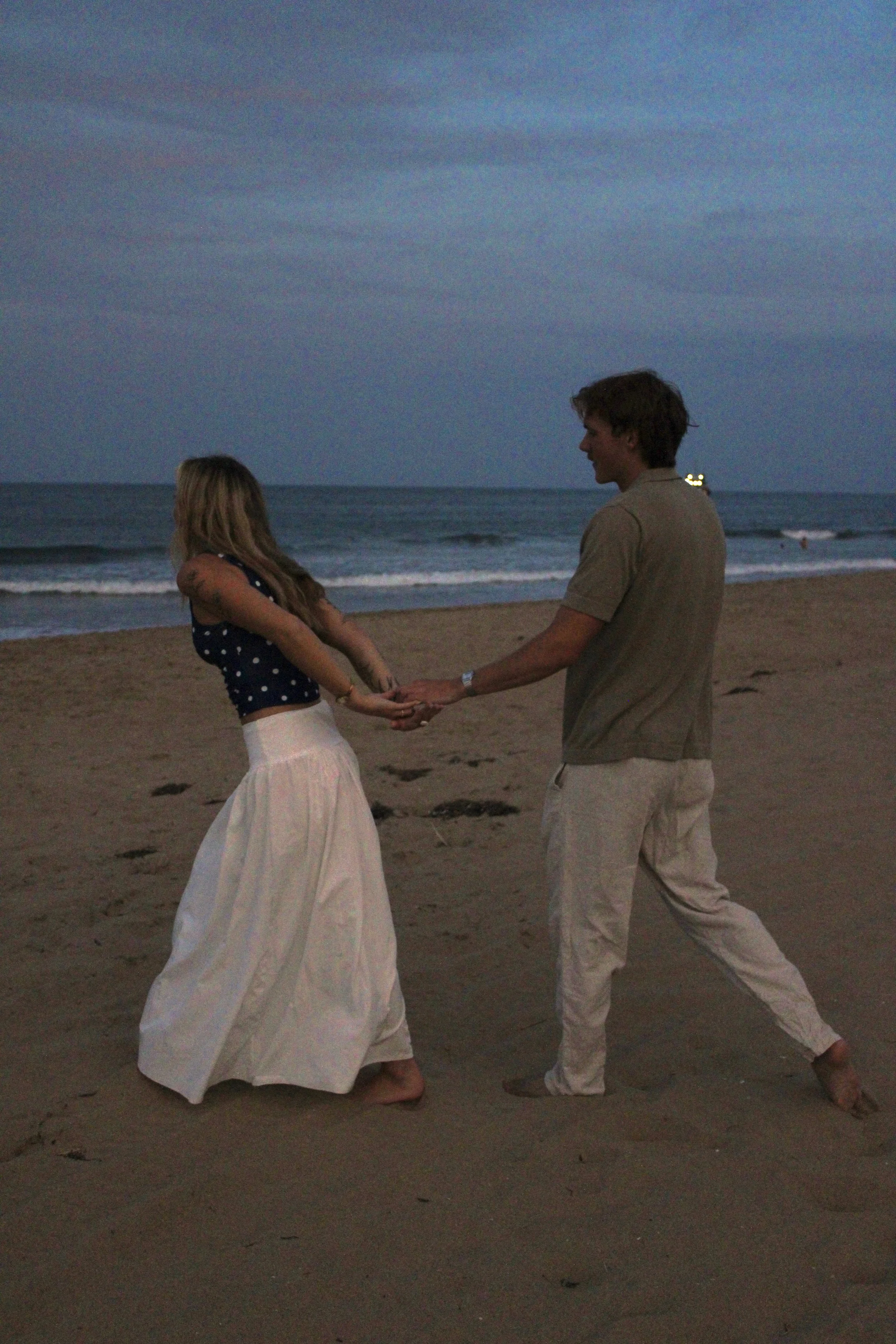 A young couple holding hands and dancing on the beach at dusk, with waves and a cloudy sky in the background.