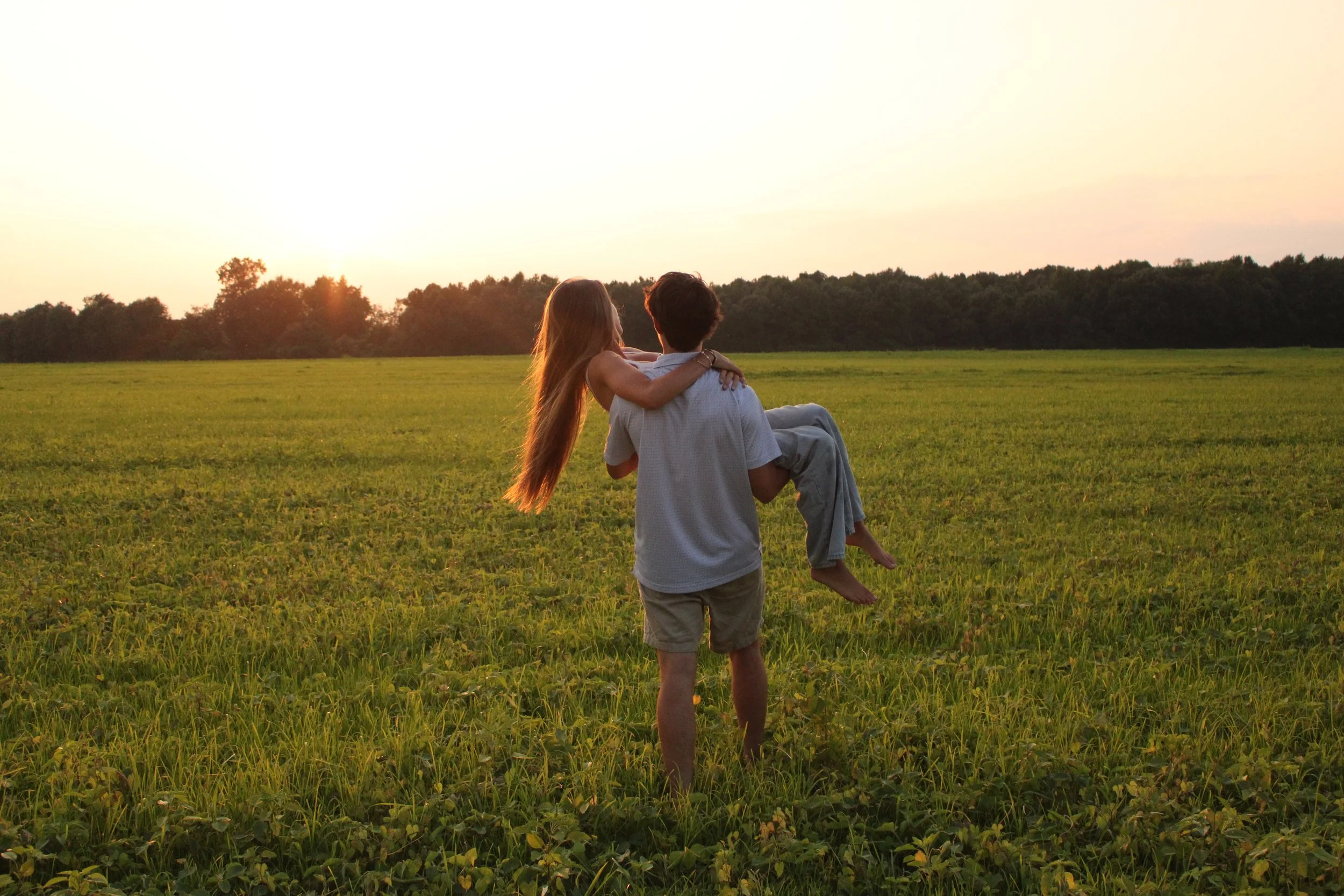 A man carrying a woman in his arms in a grassy field at sunset, with a line of trees in the background.