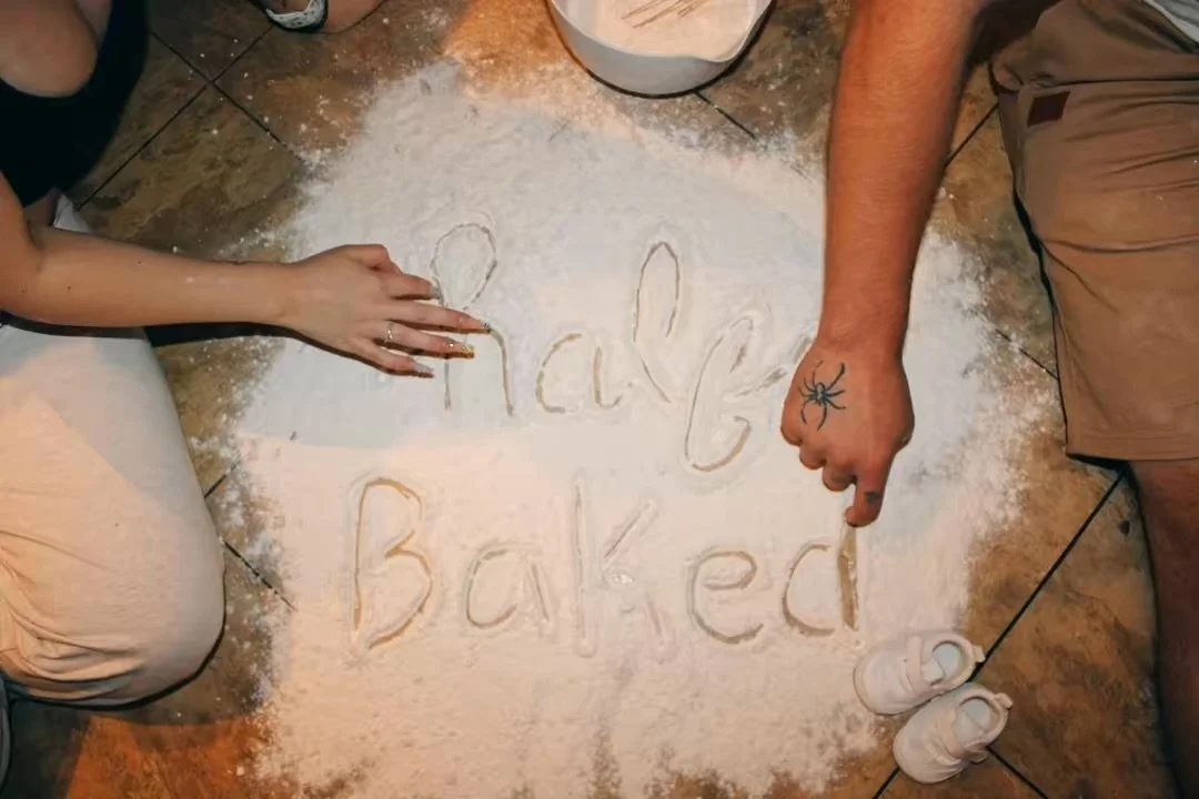 A person is writing 'Hello Baked' on a flour-covered floor with two fingers, while another person is pointing at the writing with a finger, near a bowl of flour and a pair of white shoes.
