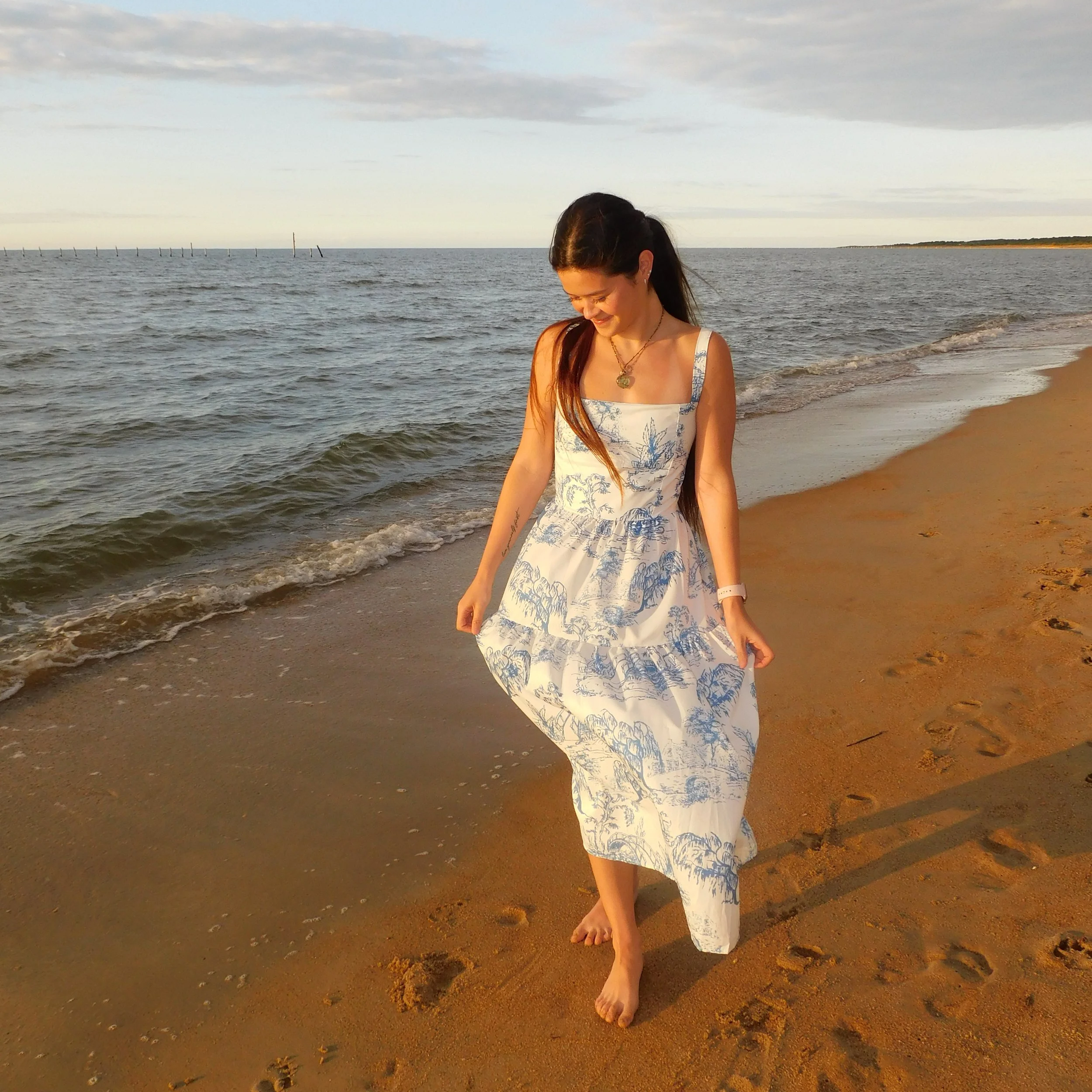 A woman with long dark hair, wearing a blue and white patterned dress, smiling and looking down while walking barefoot on the beach during sunset, with the ocean and cloudy sky in the background.