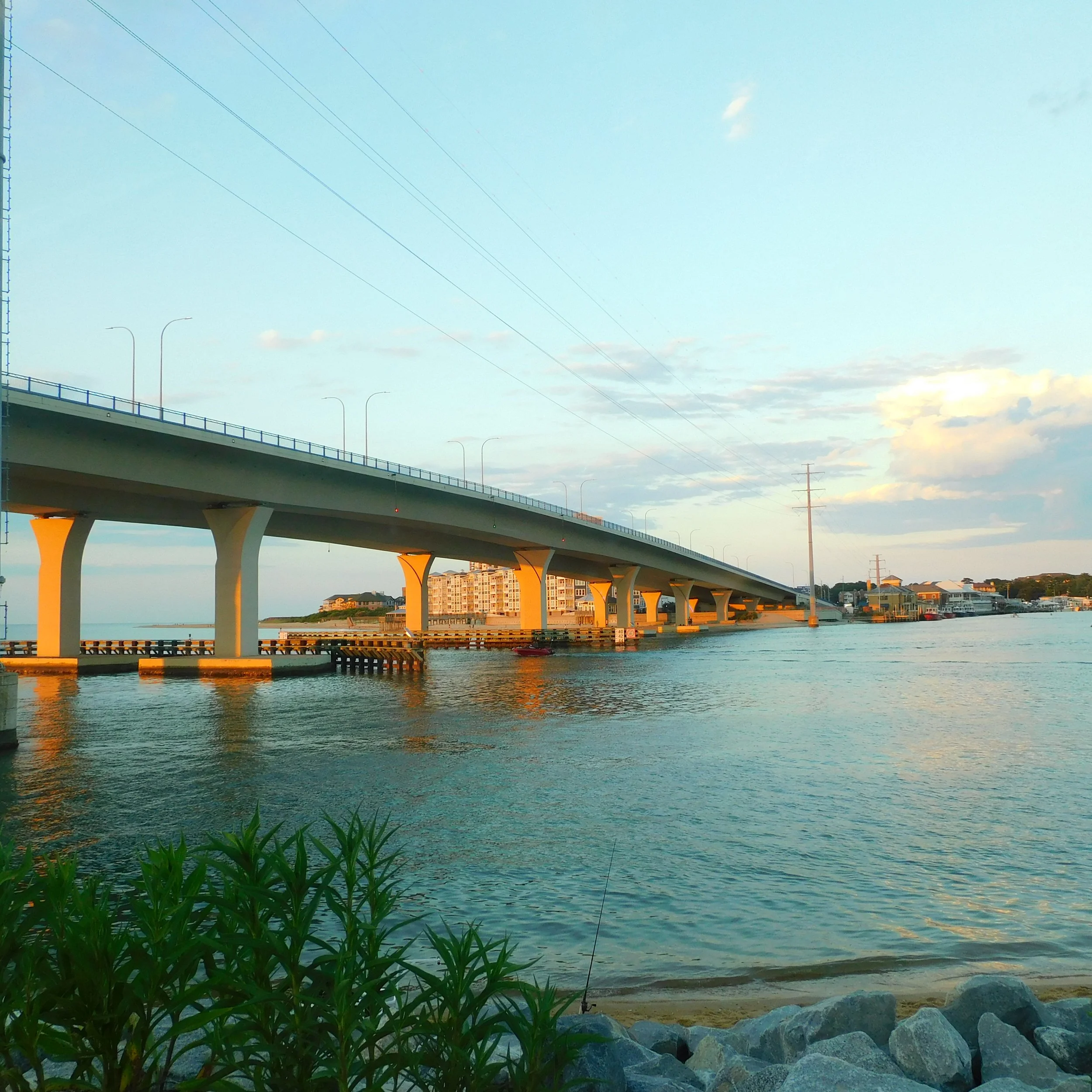 A bridge over a body of water with houses and trees in the background, and plants and rocks in the foreground, with a blue sky and clouds above.