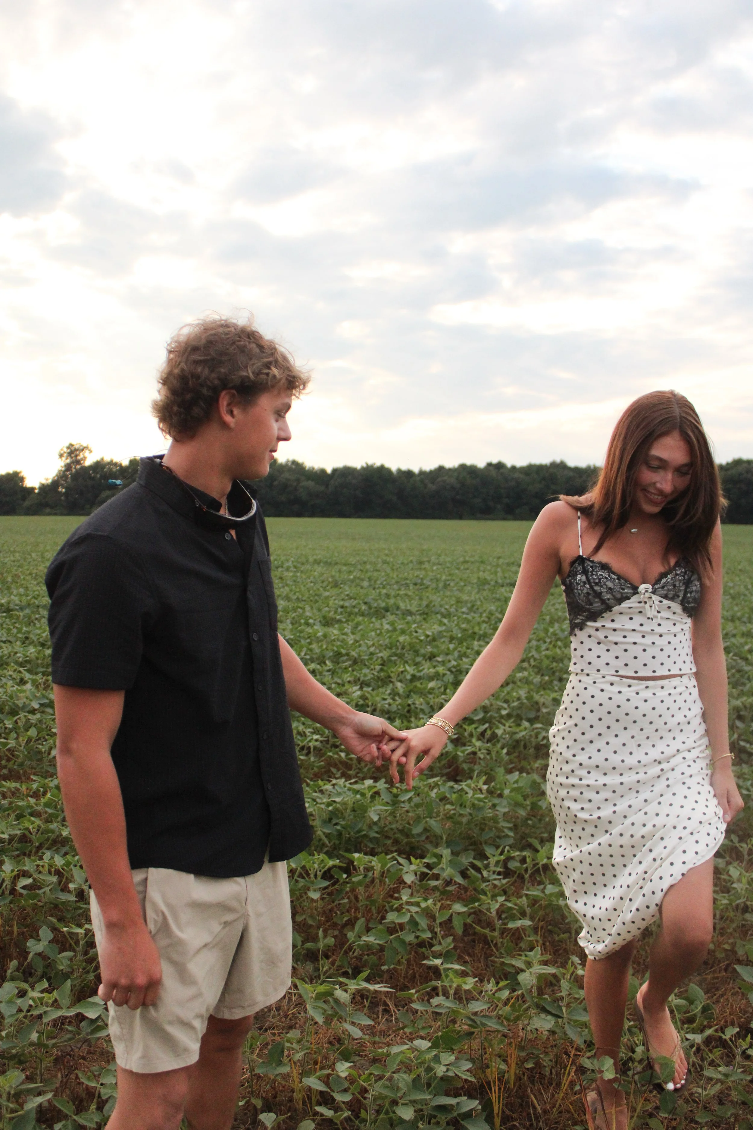 A man and a woman holding hands and walking through a green field on a cloudy day.