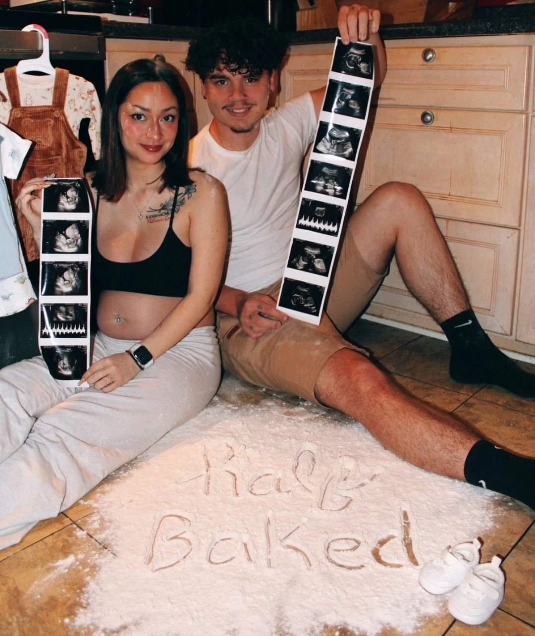 A couple sitting on the kitchen floor, holding ultrasound pictures, with the words 'thely baked' written in flour on the floor, suggesting an upcoming baby.