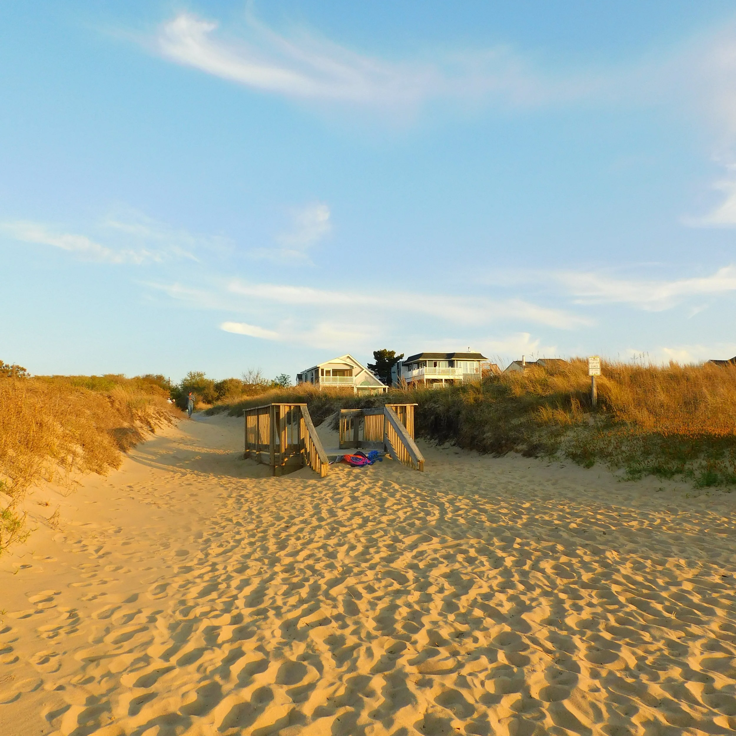 A sandy beach pathway with footprints, leading to houses on the sand dunes in the distance, under a blue sky with some clouds.