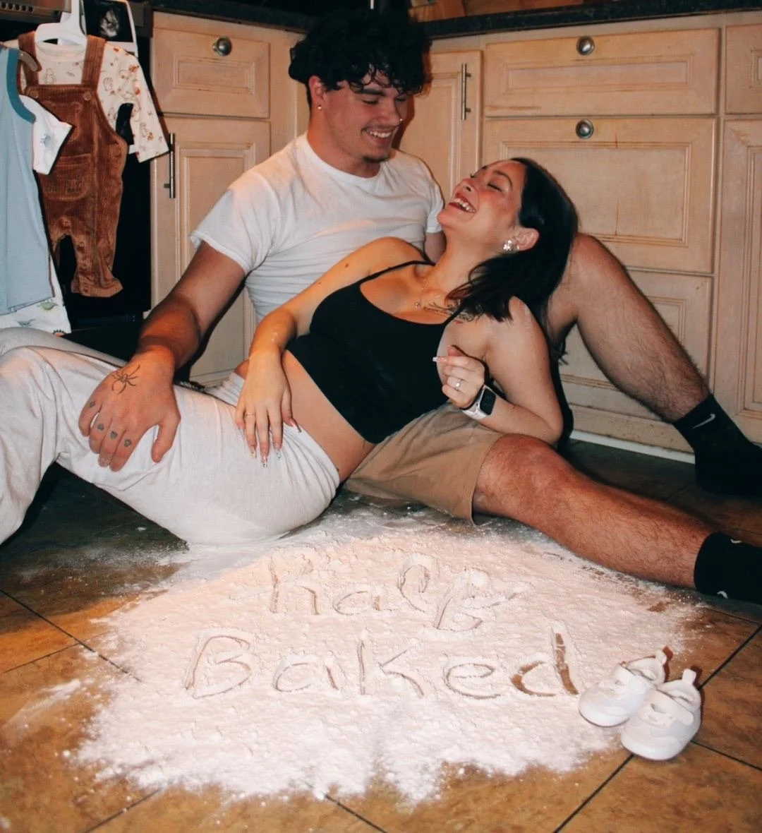 A couple sitting on the floor in a kitchen, smiling and laughing. The woman is lying back on the man's lap, and they are surrounded by flour with the phrase 'We Banged' written in it. A pair of white baby shoes is next to the writing.