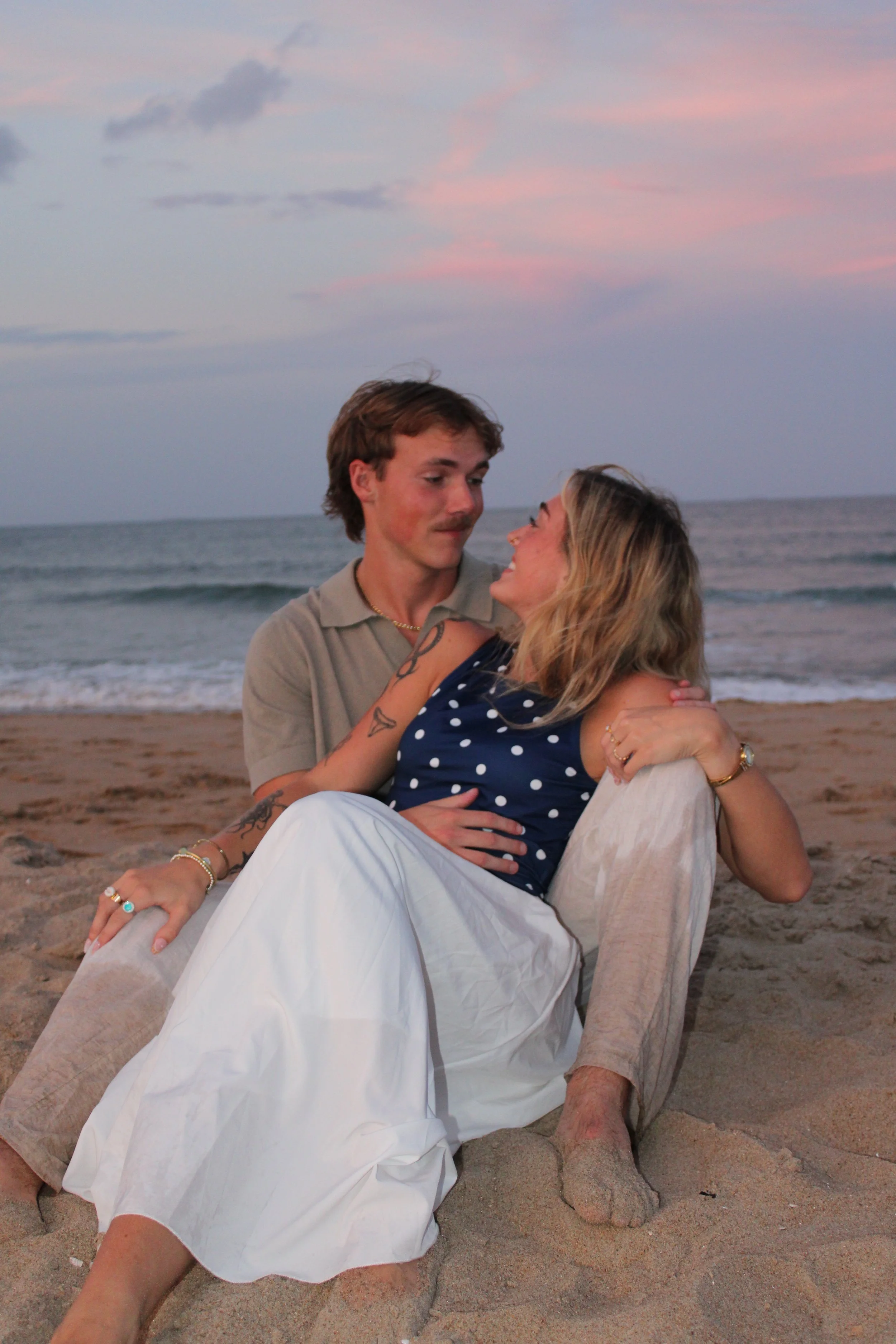 A couple sitting on the sandy beach at sunset, looking at each other, with ocean waves in the background and pink skies overhead.