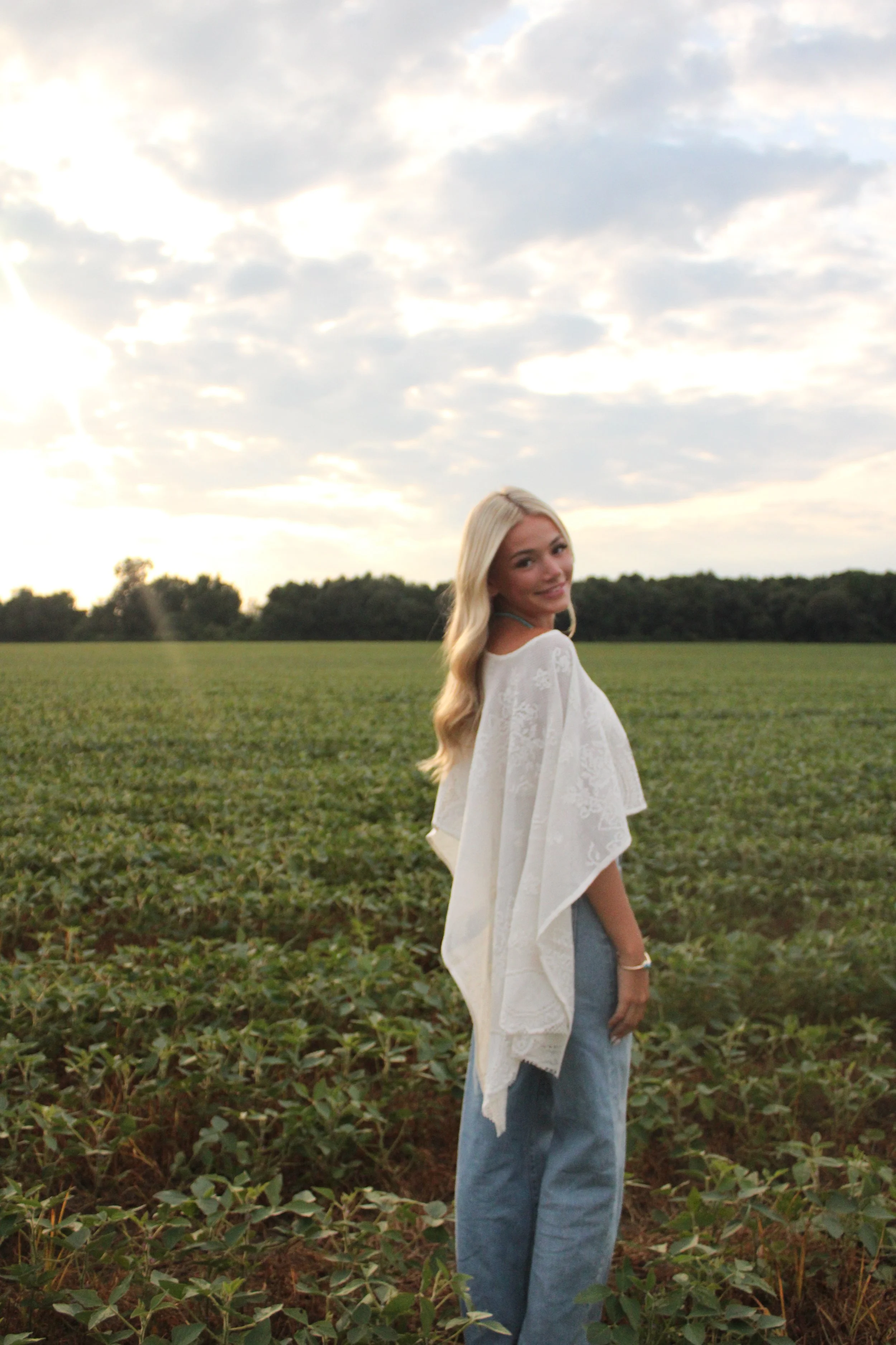 A young woman with long blonde hair standing in a green field under a cloudy sky during sunset, wearing a white top and blue jeans, smiling while looking back at the camera.