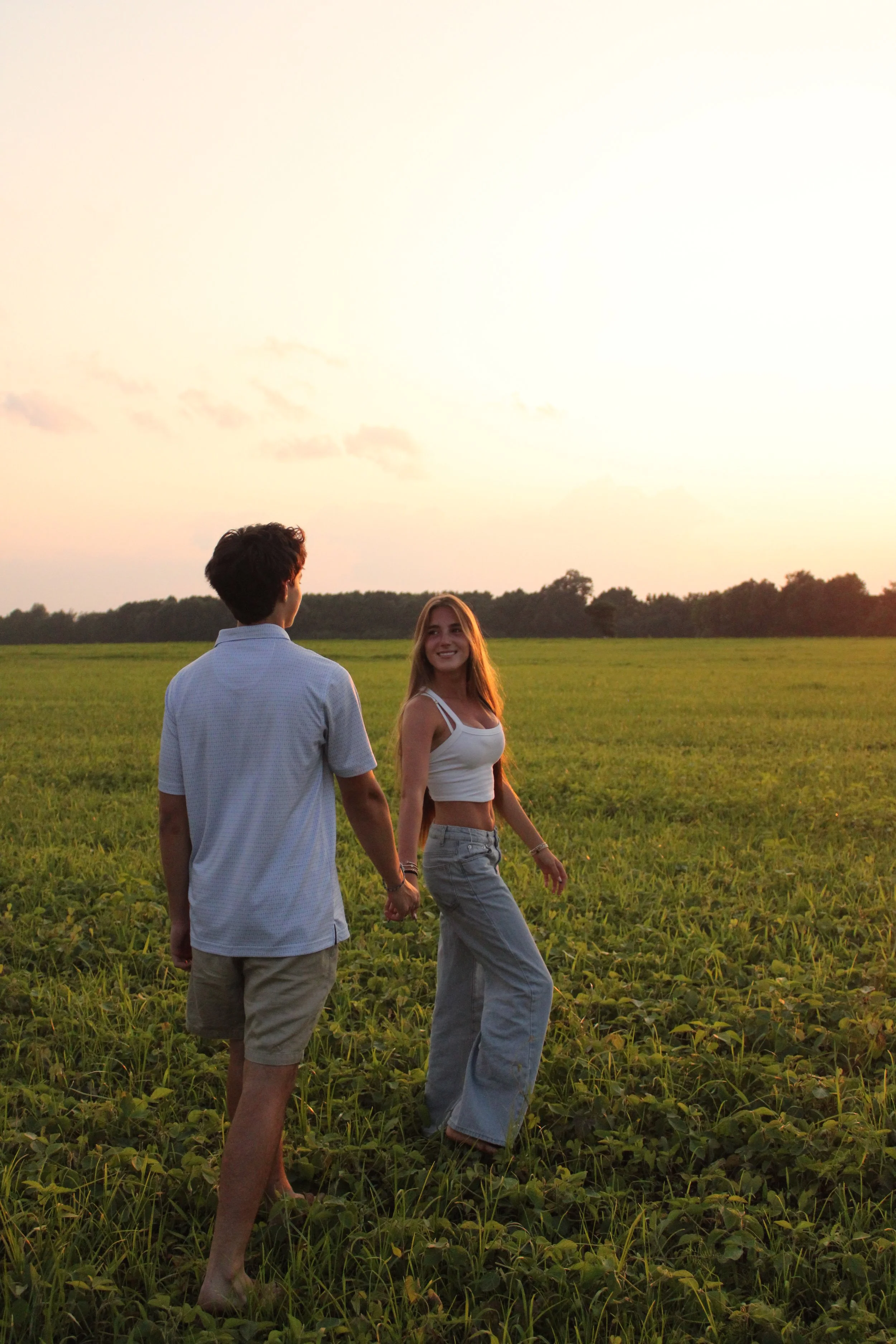 A couple holding hands and walking in a green field during sunset, with a man facing away and a woman smiling at him.