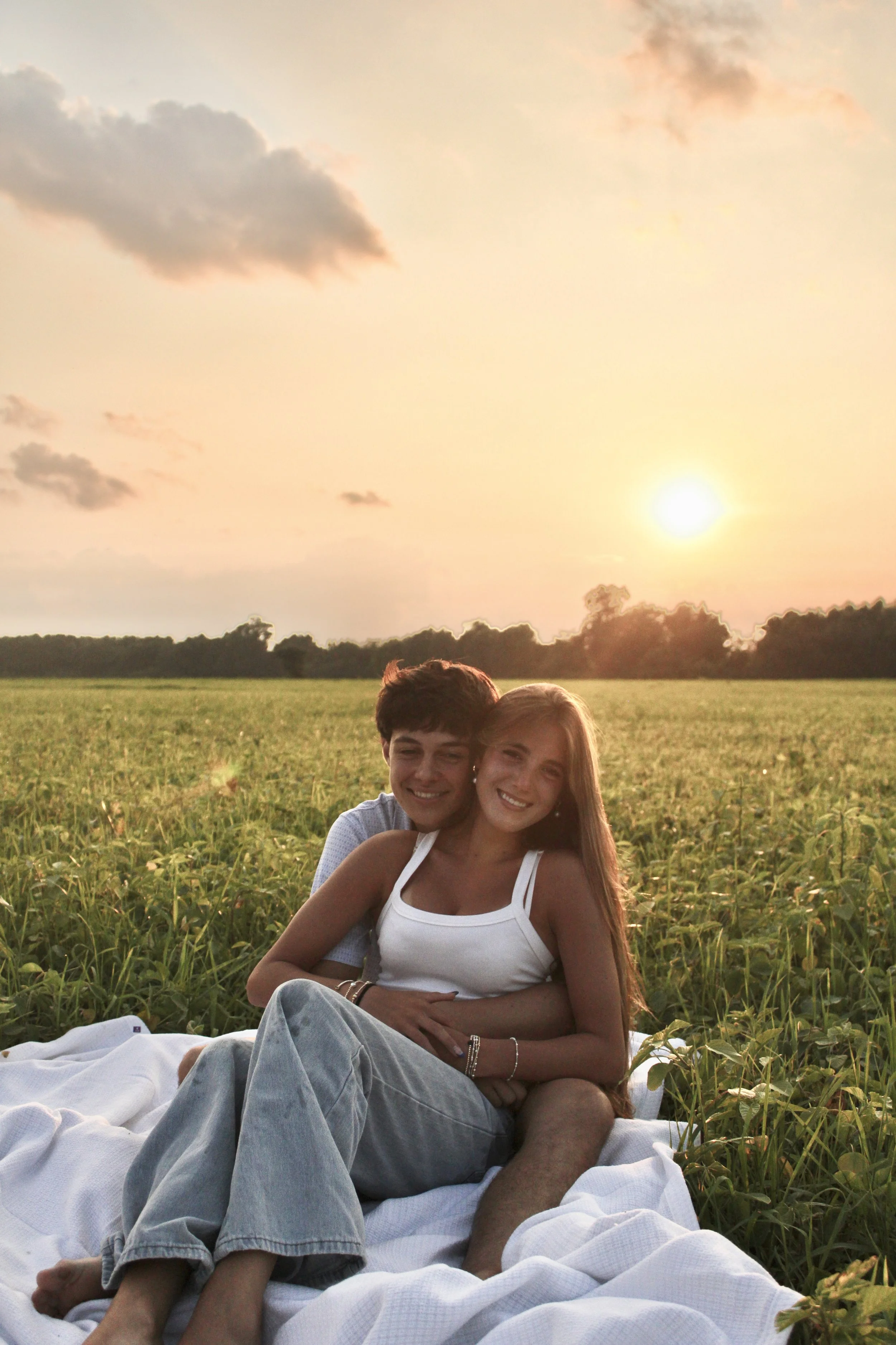 A young man and woman sitting together on a blanket in a green field at sunset, smiling and embracing each other.