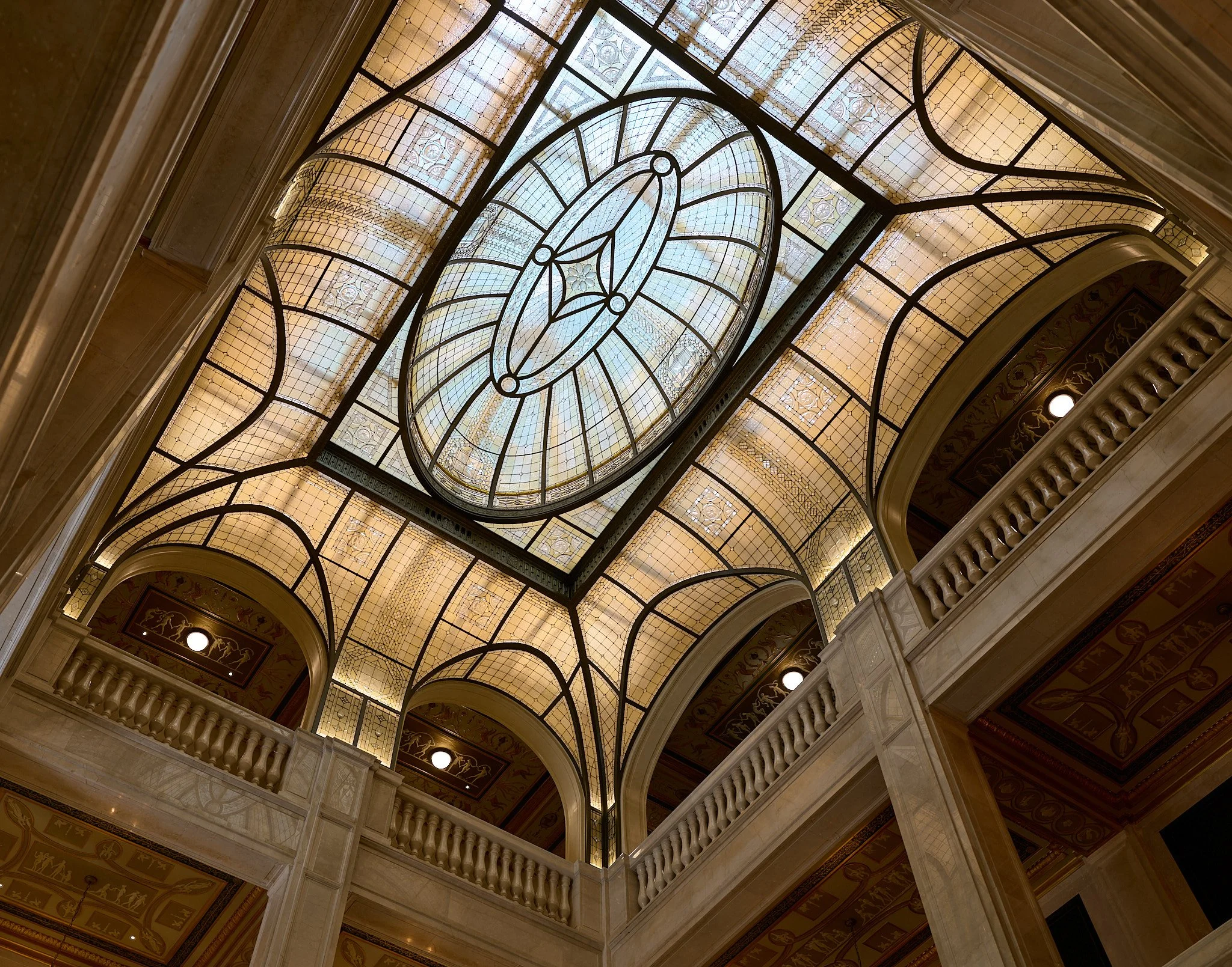 Interior view of a grand building with a stained glass ceiling, ornate railings, and decorative arches.