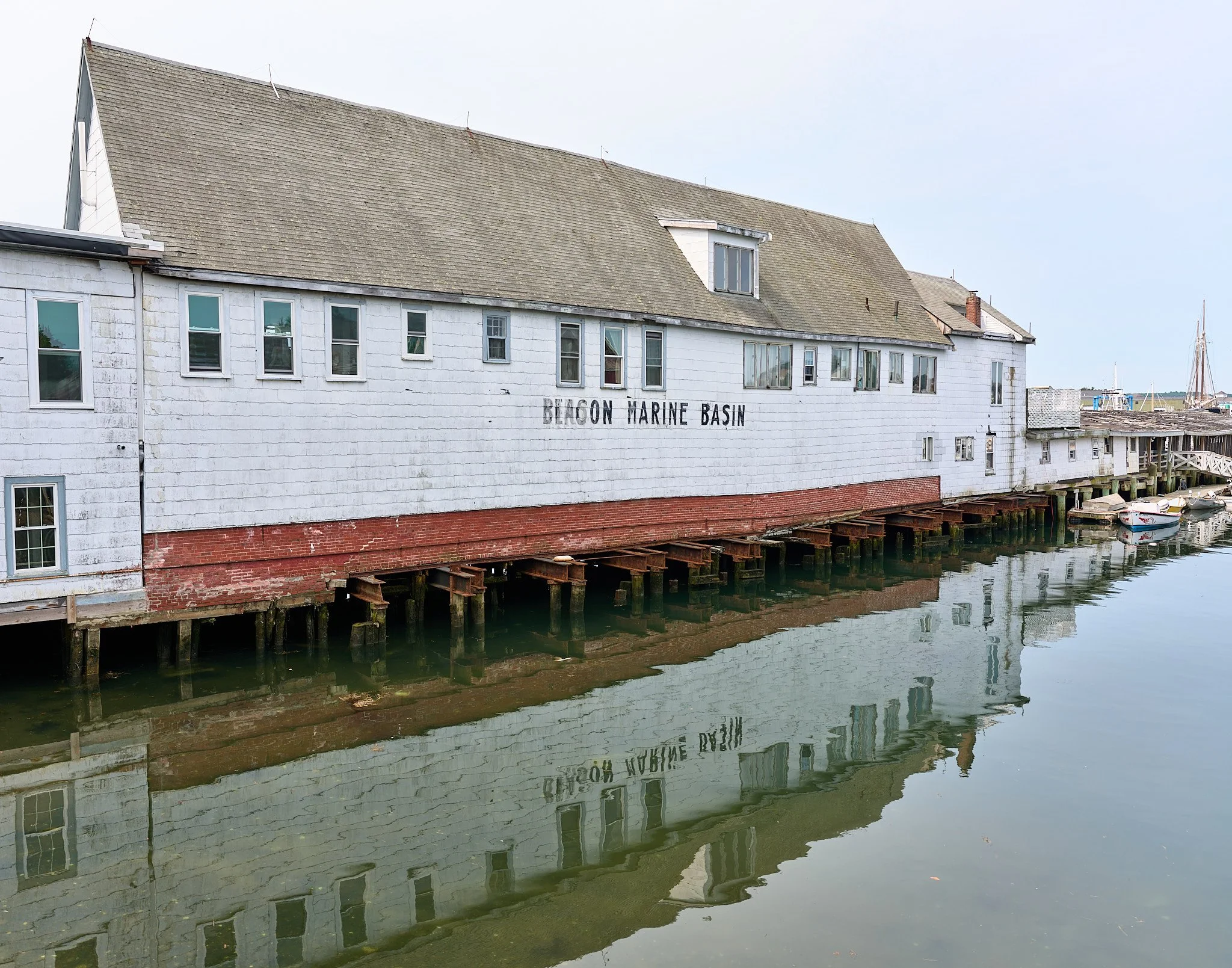 A white building with the words 'BEACON MARINE BASIN' on its side, built on stilts over a body of water, with boats and sailboats in the background.