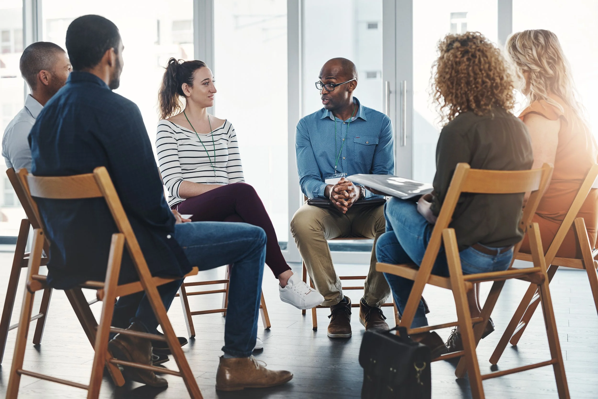 A diverse group of six adults sitting in a circle in a bright office, engaged in a discussion, with a man speaking to the group.
