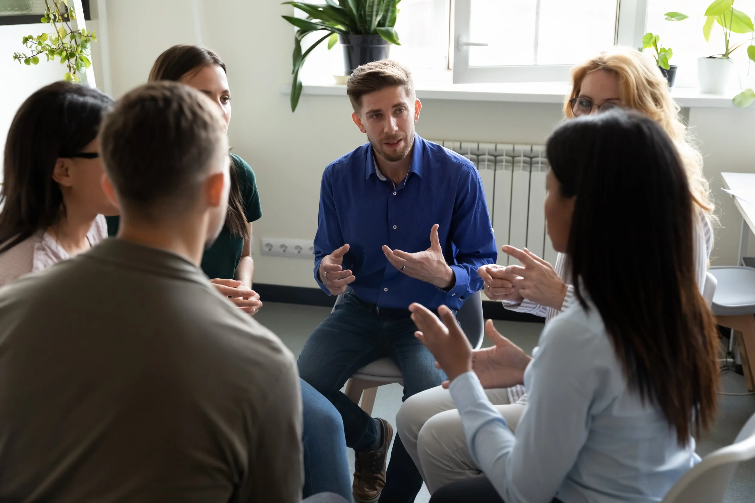 A group of six people sitting in a circle having a discussion, with a young man in a blue shirt talking and gesturing with his hands. The room has potted plants on the windowsill and natural light coming through the windows.