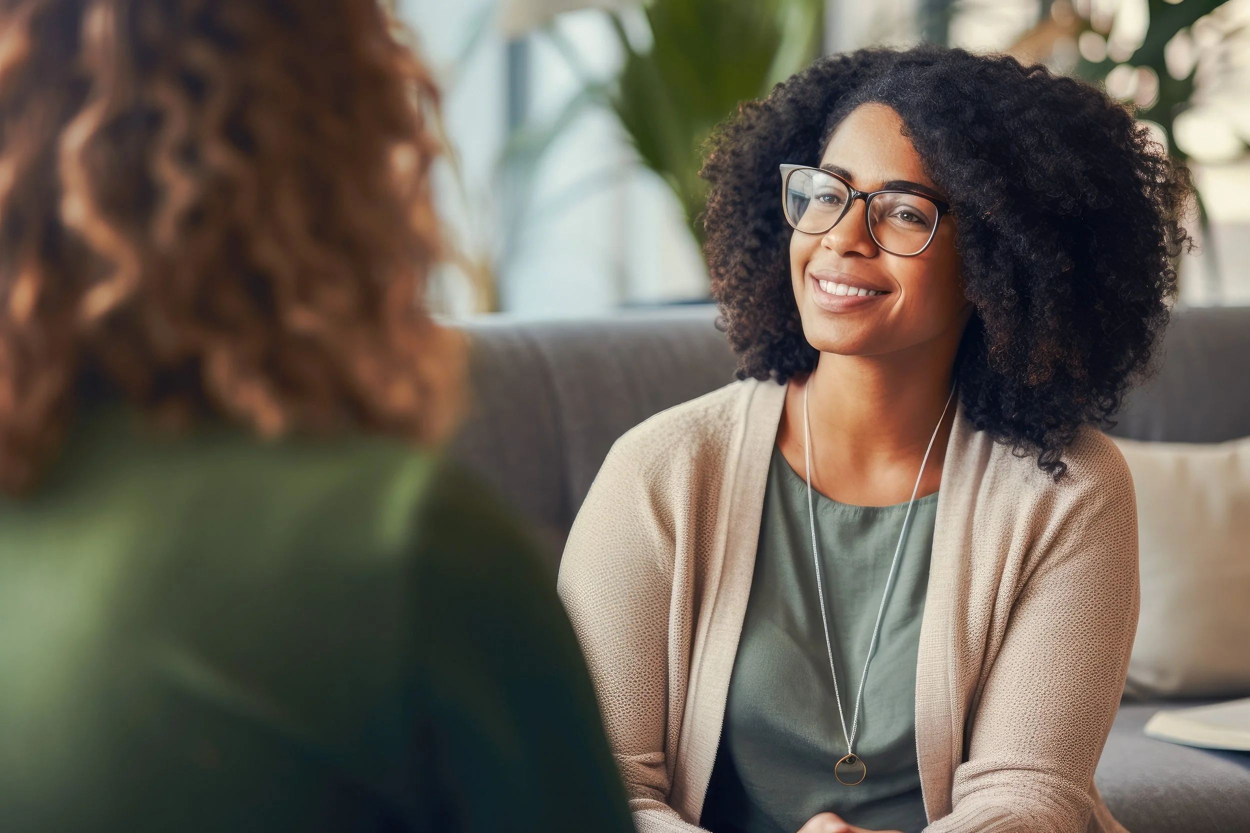 A woman with curly black hair, glasses, and a light-colored blazer smiling and talking to another person with curly hair, in an indoor setting with plants in the background.