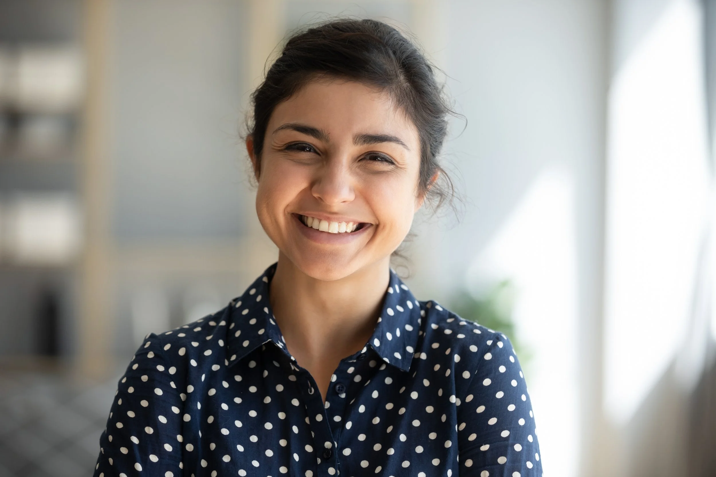 A woman smiling with short dark hair, wearing a navy blue polka dot shirt, in a bright indoor setting.