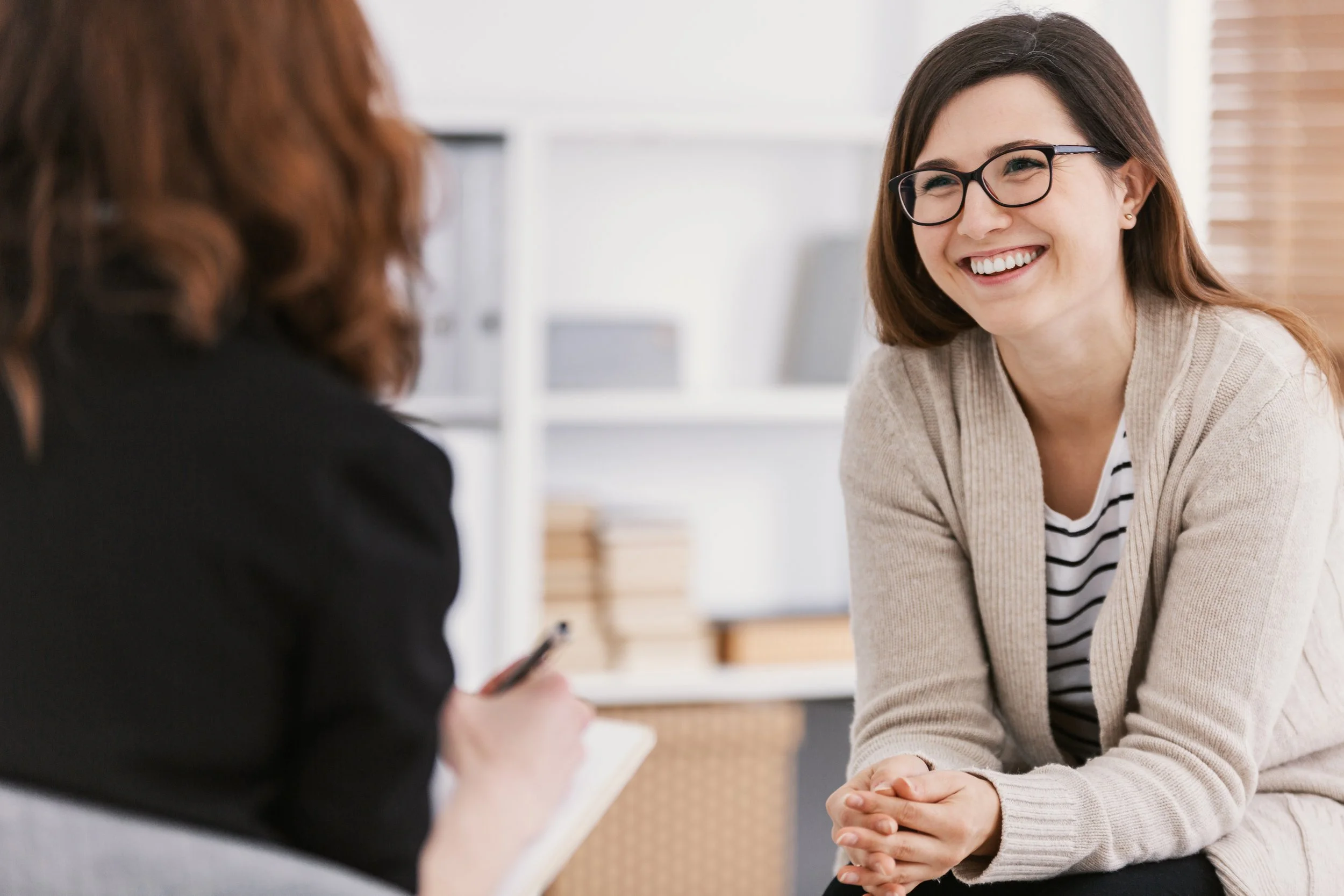A woman wearing glasses smiling and engaging in conversation with another person in an indoor setting.