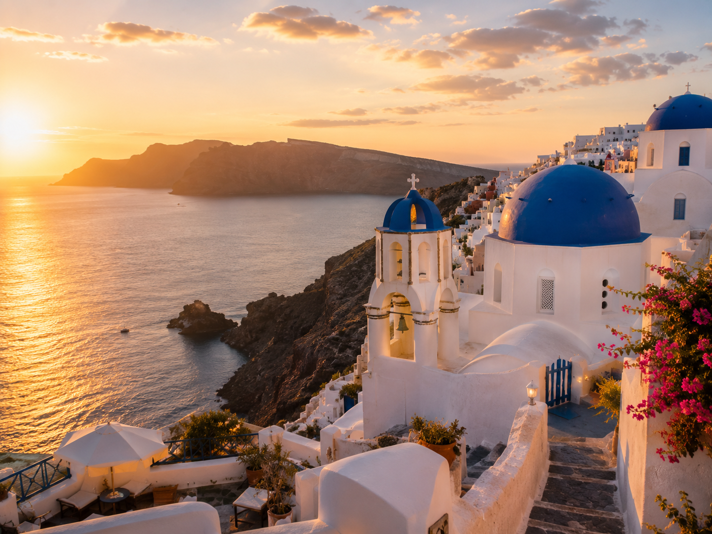Sunset over the Aegean Sea in Santorini, Greece, with iconic white buildings and blue domes, a bell tower, and vibrant flowers on the hillside.