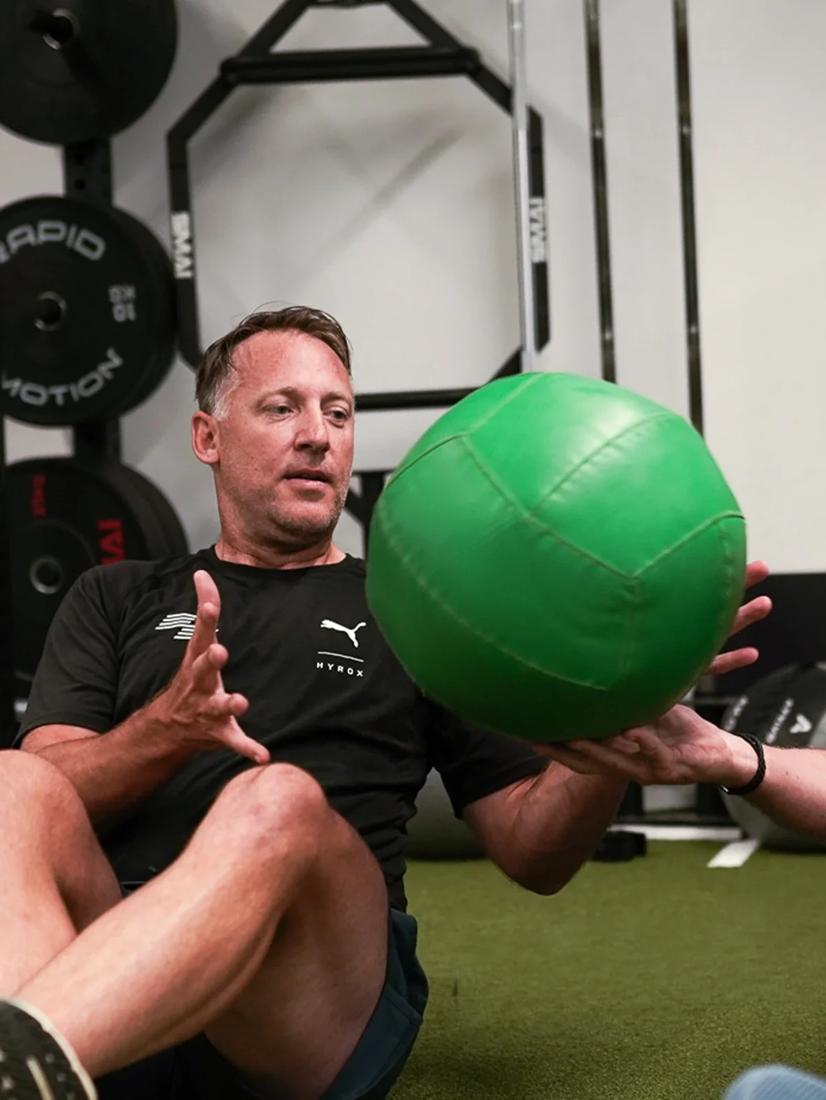 A man sitting on a gym floor holding a large green exercise ball with another person's hand supporting from underneath. The man is wearing a black Puma shirt with
