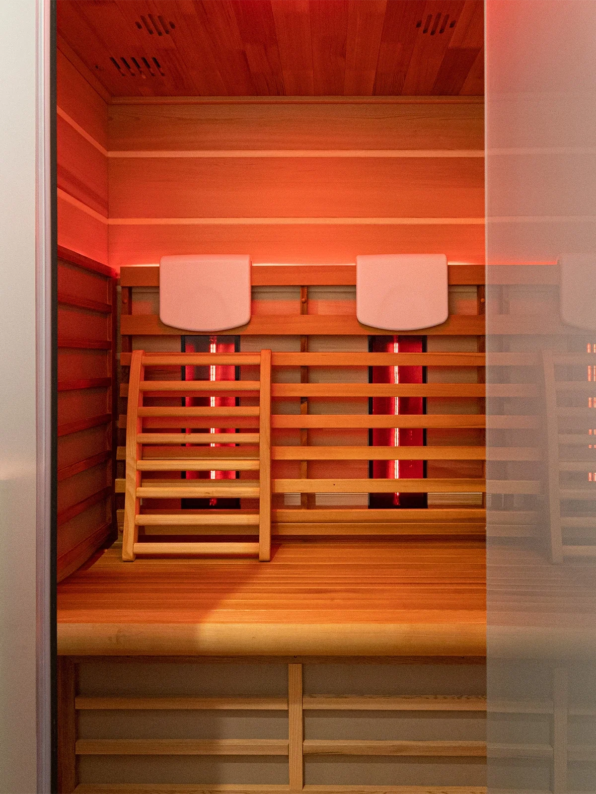 An interior view of a traditional Finnish sauna made of light-colored wood, featuring a ladder, two white headrests, and infrared heaters on the wall.