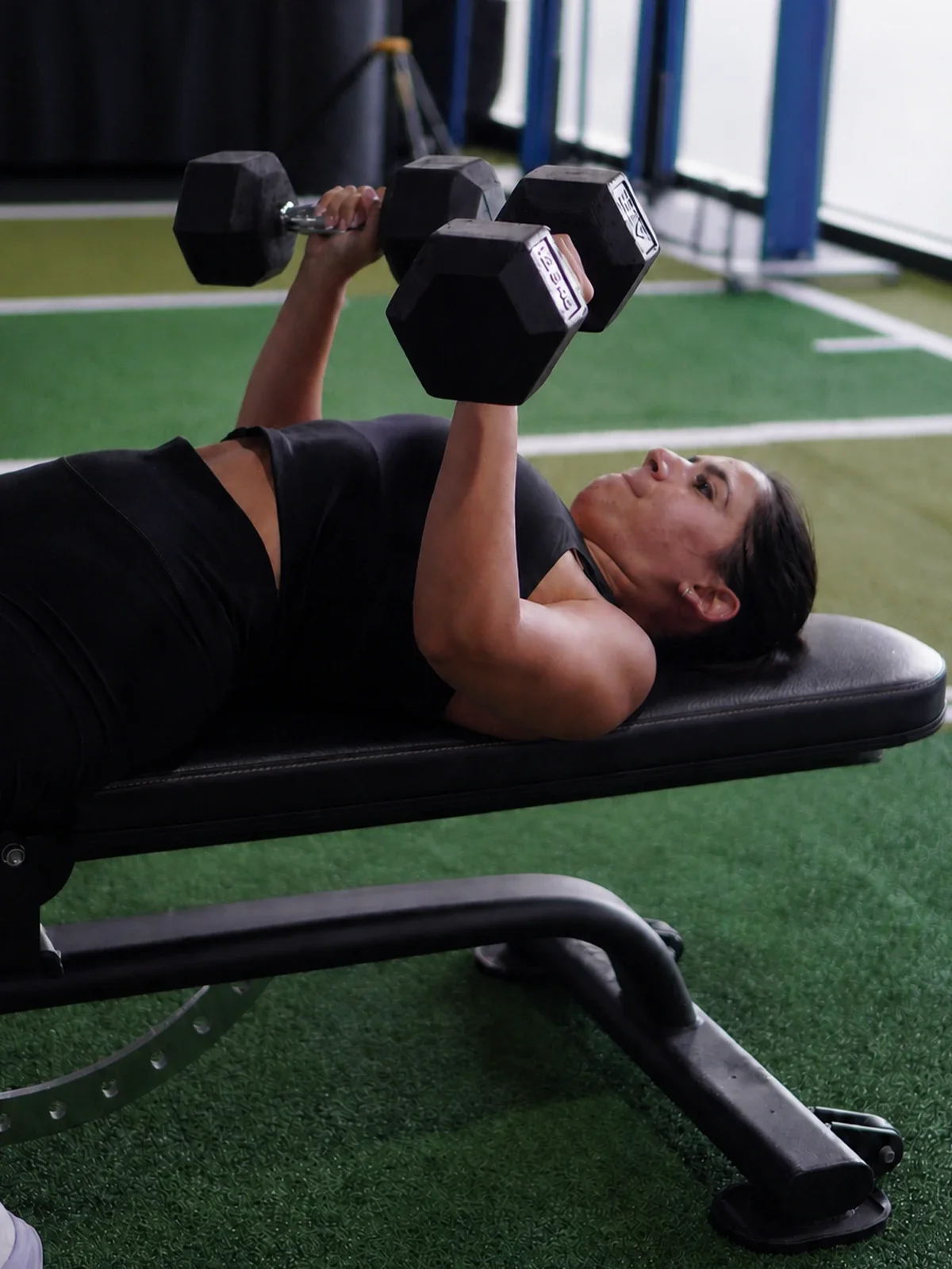 A woman lies on a black workout bench and lifts two black dumbbells while exercising in a gym with artificial turf flooring.