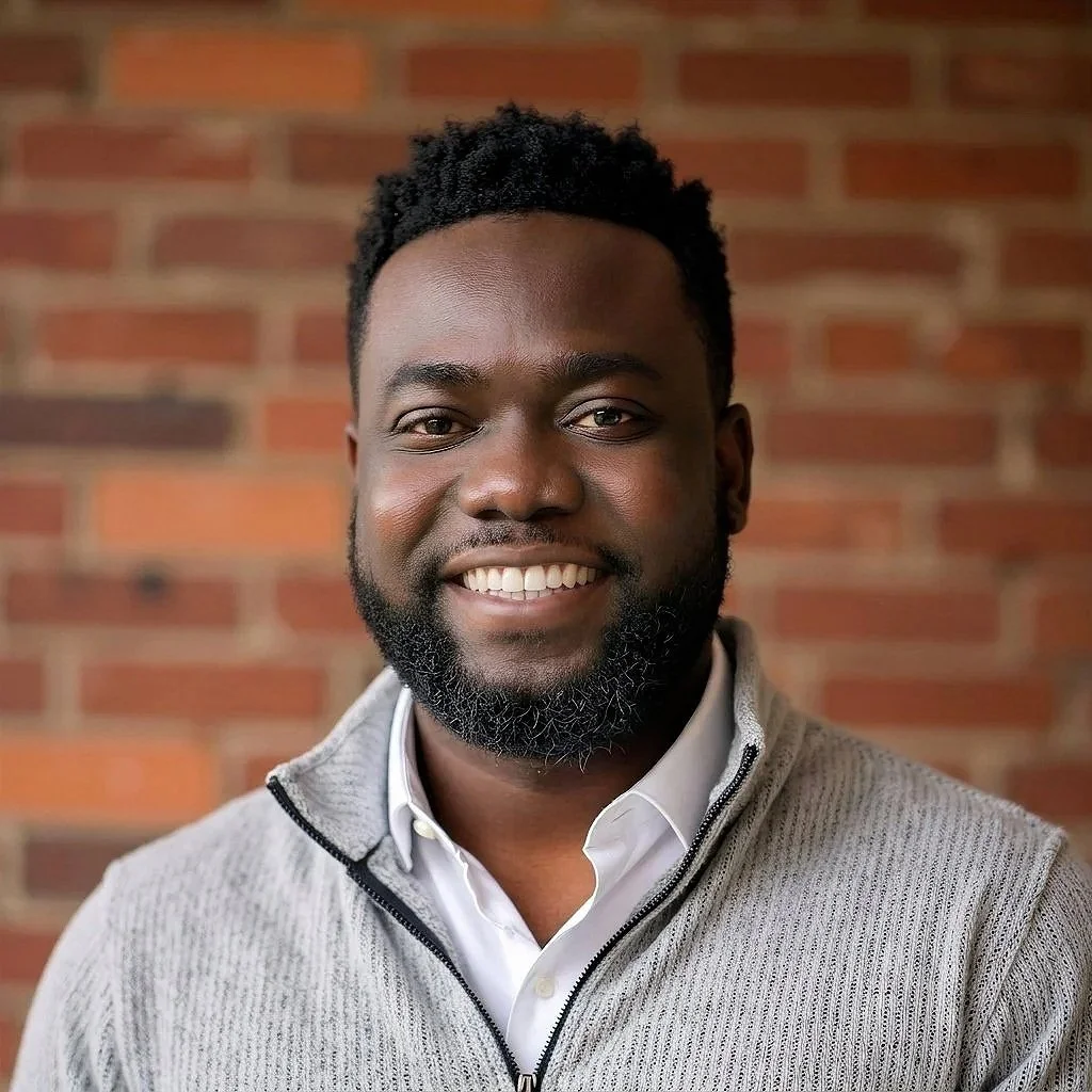 A smiling Black man with a beard, wearing a white shirt and a light gray zip-up sweater, standing in front of a brick wall.