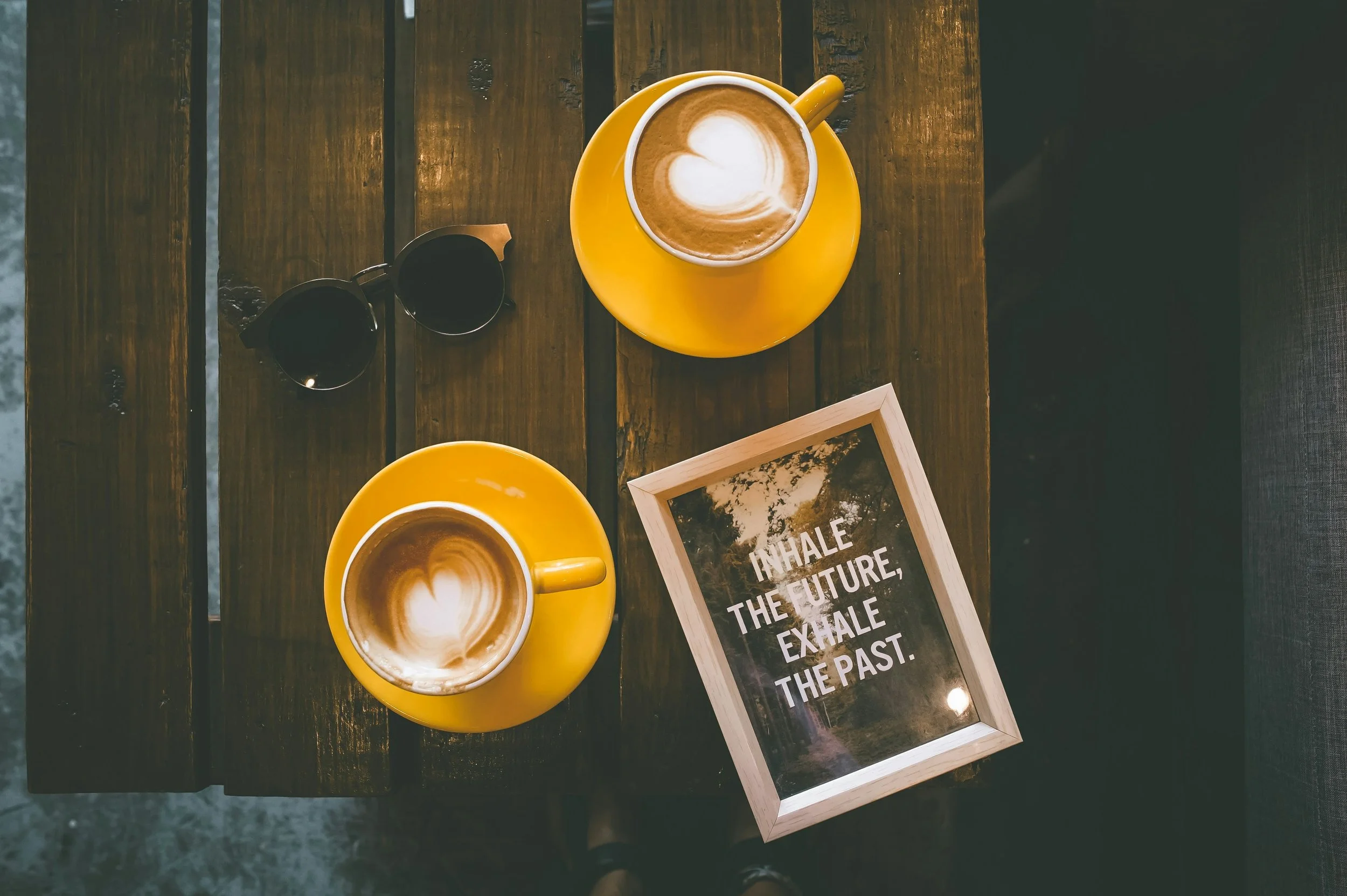 A top-down view of two yellow cups of coffee with latte art resembling hearts, placed on a rustic wooden table. There are also dark-colored sunglasses, and a framed quote that reads 'INHALE THE FUTURE EXHALE THE PAST'.