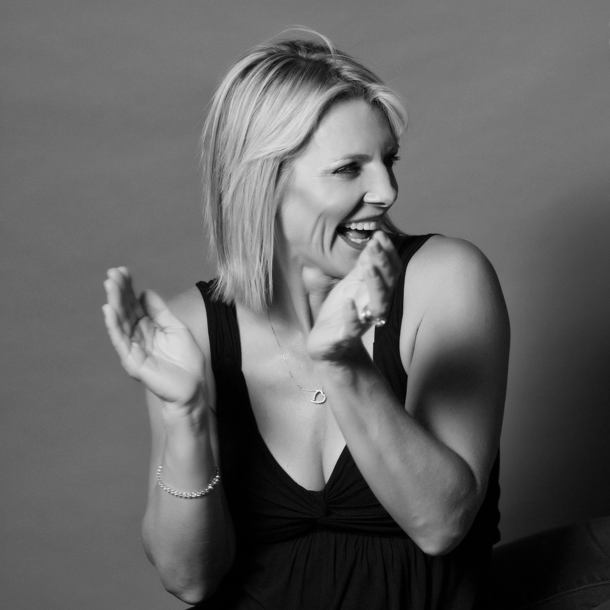 A woman with shoulder-length blonde hair smiling and laughing, wearing a black sleeveless top, a pearl bracelet, and a necklace, against a plain background.