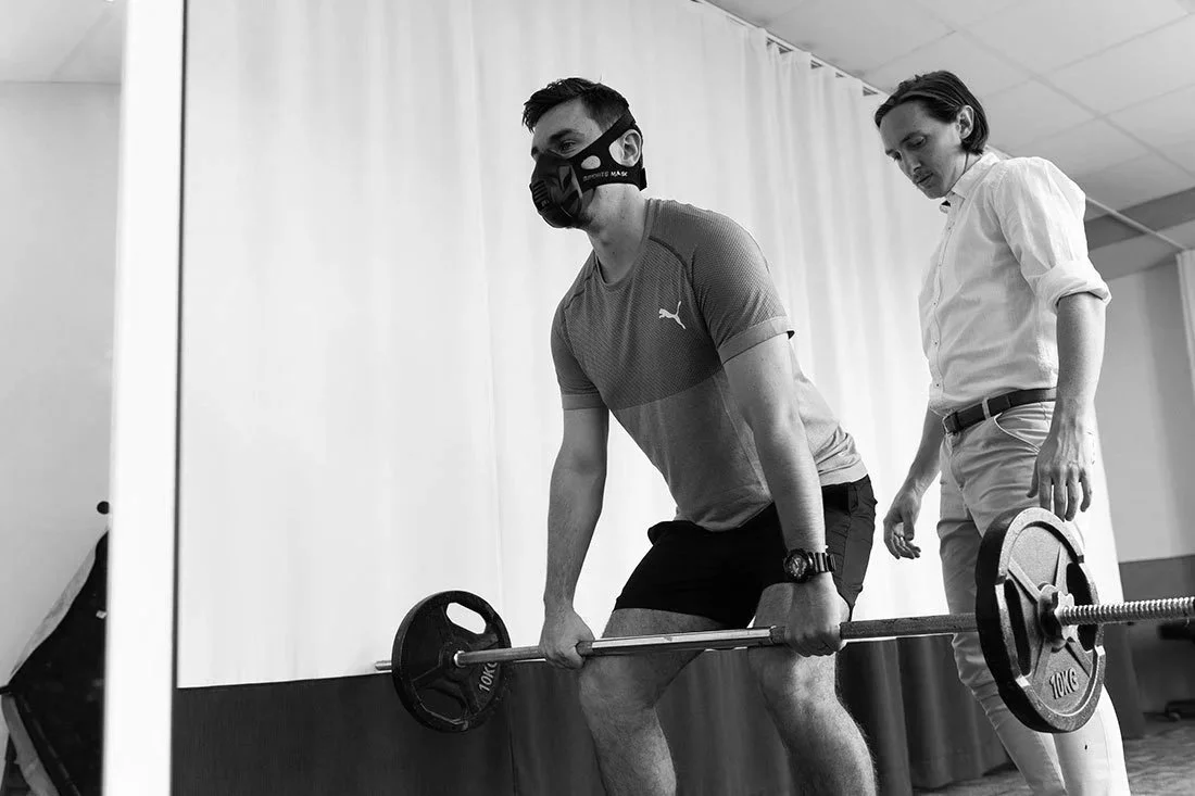 A man wearing a face mask lifting a barbell with weights during a workout session, guided by a woman in a white shirt in a gym or fitness facility.