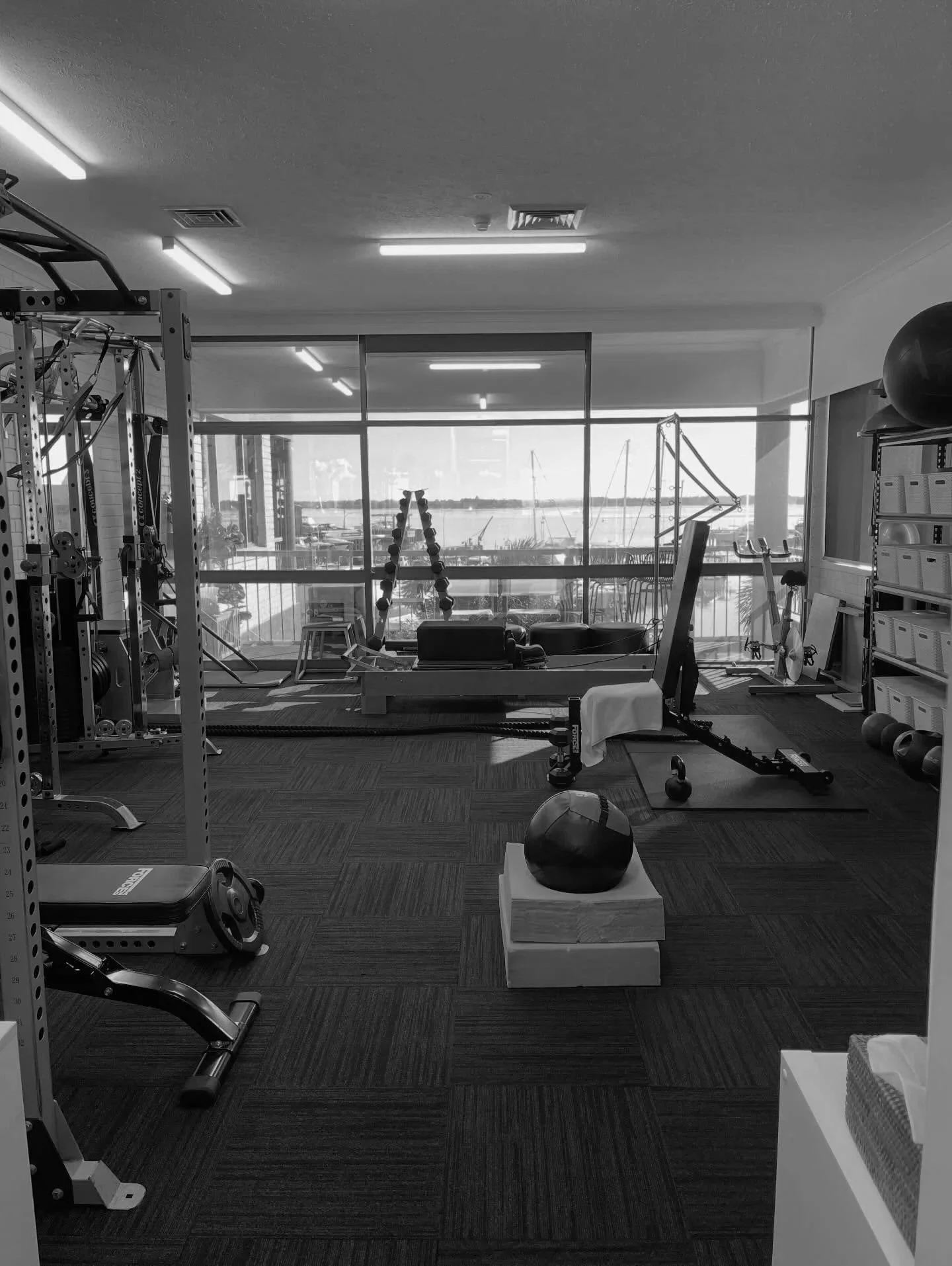 Empty fitness gym with various workout equipment including a squat rack, kettlebell, exercise bike, stability ball, and foam roller, with large windows overlooking a waterfront view.