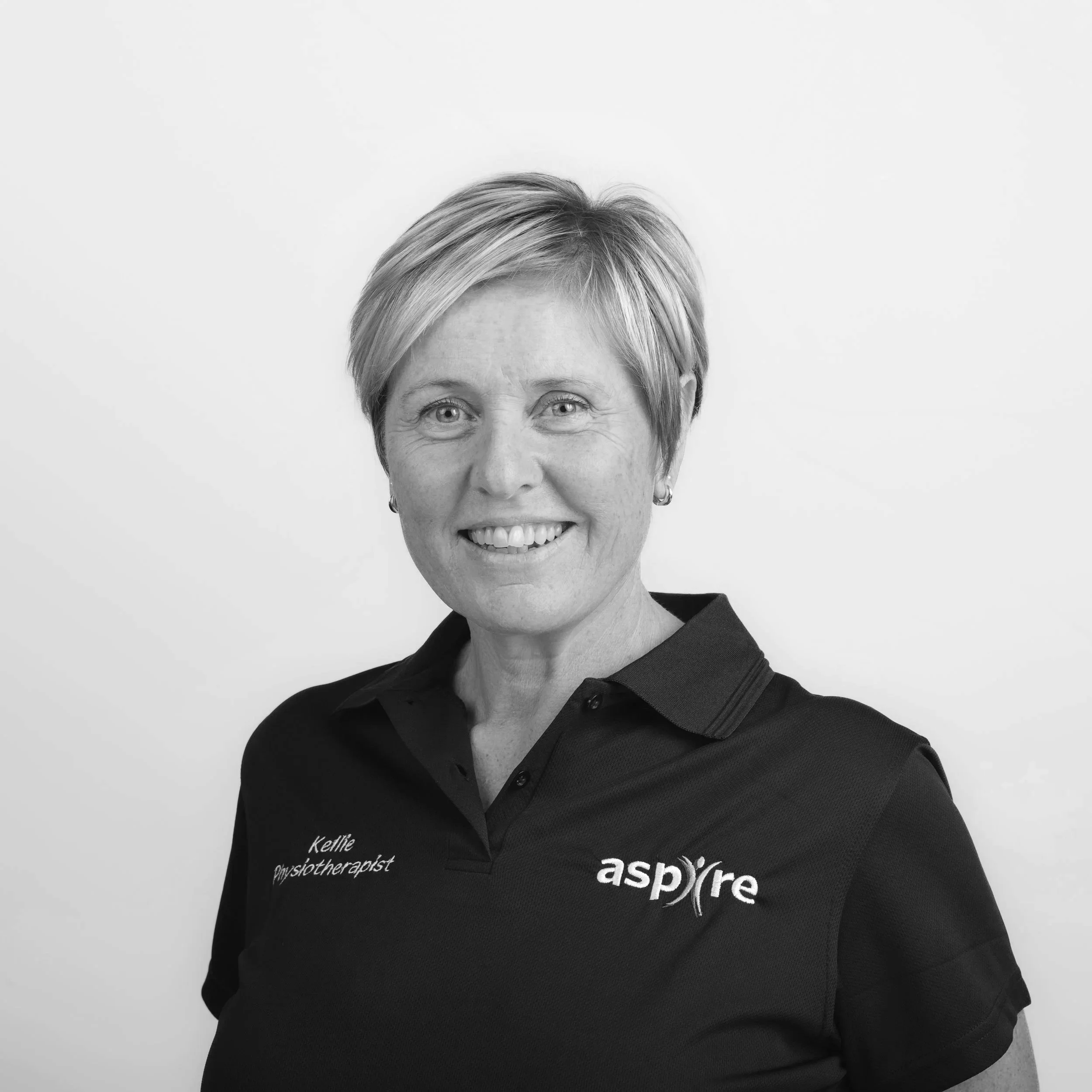 Close-up of a smiling woman with short hair wearing a black polo shirt with logos and text, against a plain white background.