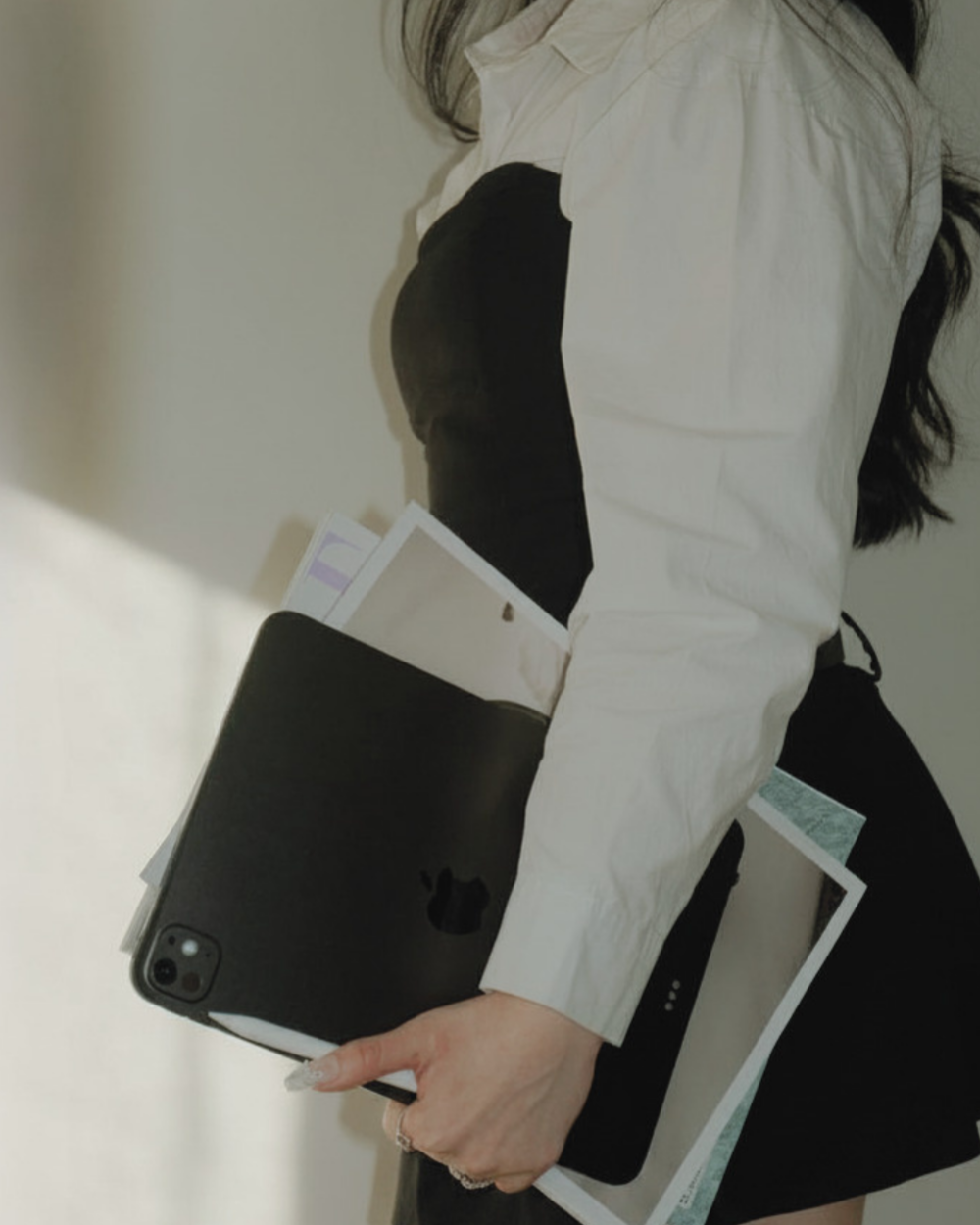 A person holding a black iPhone with a black case, a newspaper, and some documents, all in one hand, while wearing a white shirt and black pants.