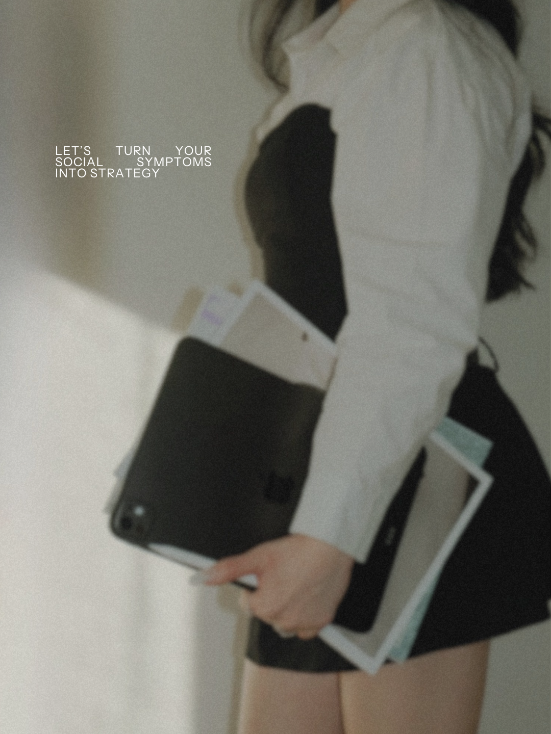 A person taking a mirror selfie, wearing a gray jacket with black detailing, holding a smartphone and some documents. Text on the image says, "Let's turn your social symptoms into strategy."