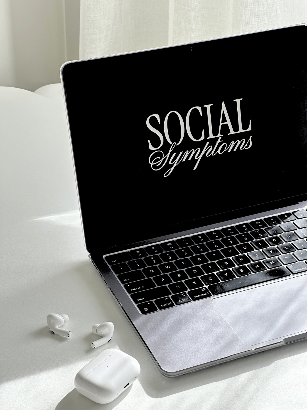Laptop on a white desk displaying the words 'Social Symptoms' on its screen, next to wireless earbuds and their charging case.