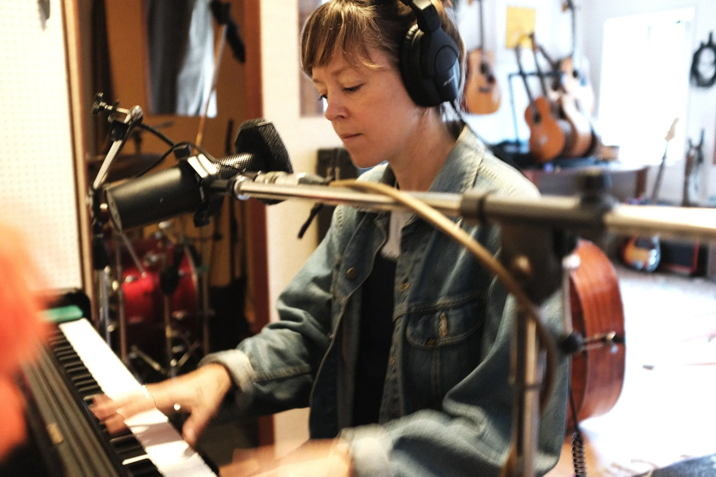 A woman with short, light brown hair wearing headphones is playing the piano in a music studio. There are guitars hanging on the wall in the background.