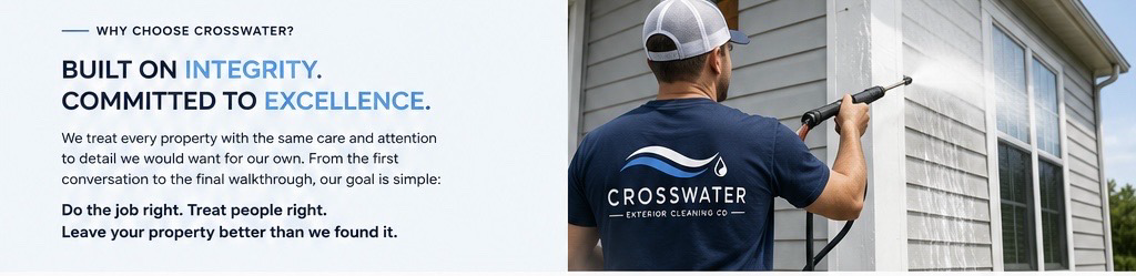 A man wearing a navy blue T-shirt with the CrossWater logo pressure washing the exterior of a house with gray siding and white trim.