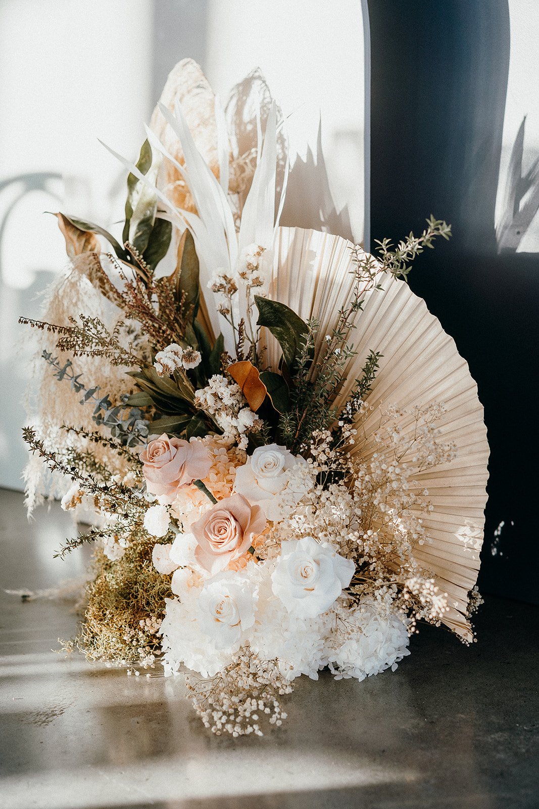 Elegant dried and fresh floral arrangement with blush roses, white flowers, greenery, and a large beige palm leaf fan