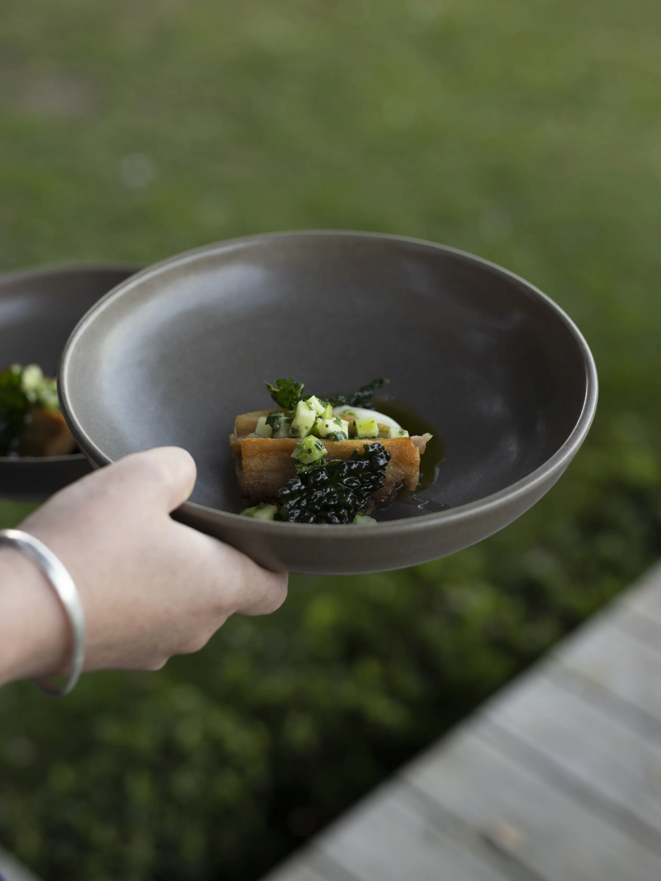 Hand holding a black bowl of gourmet food topped with diced cucumber and greens