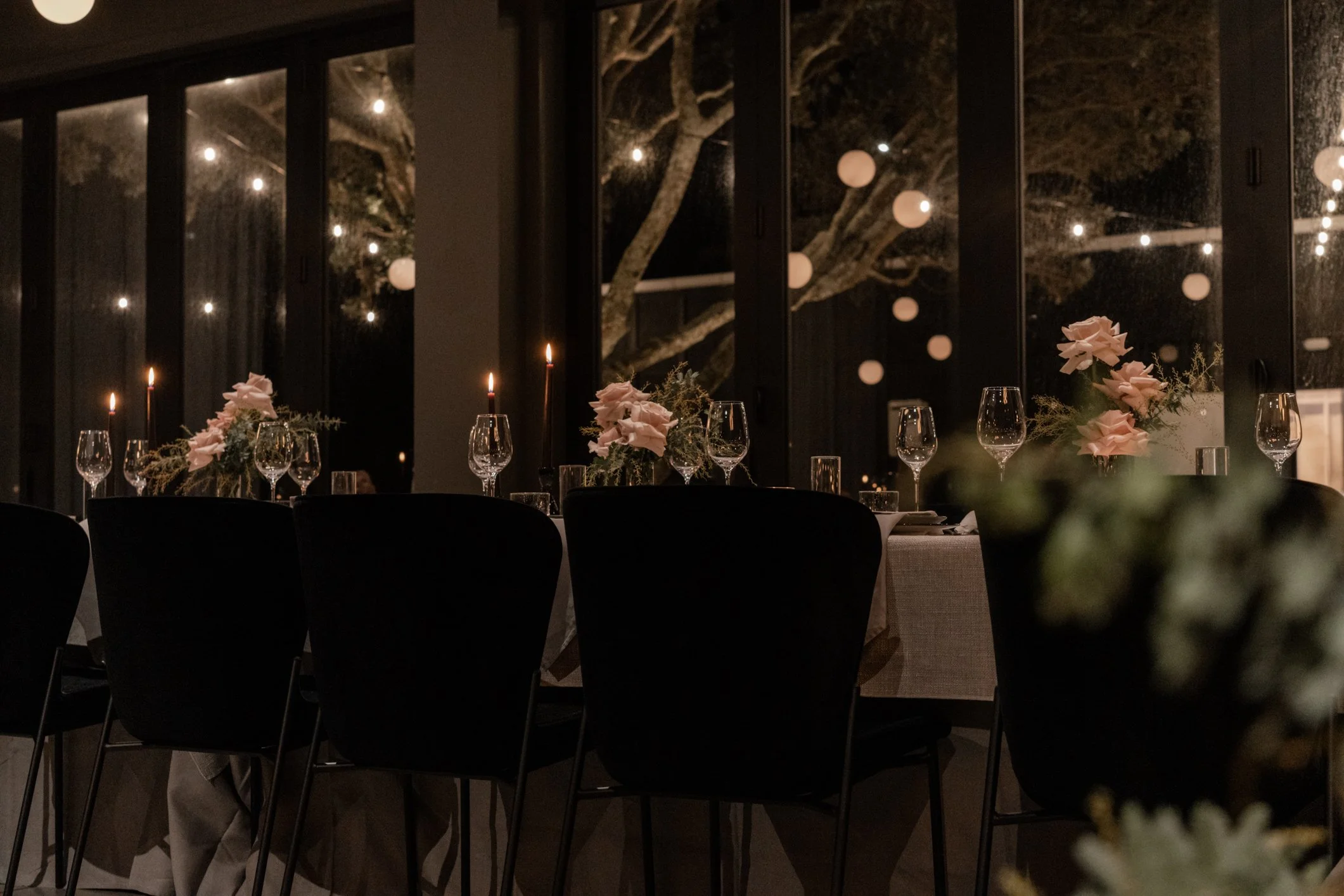 Elegant candlelit dining table with pink floral arrangements, wine glasses, dark chairs, and string lights by large windows at night