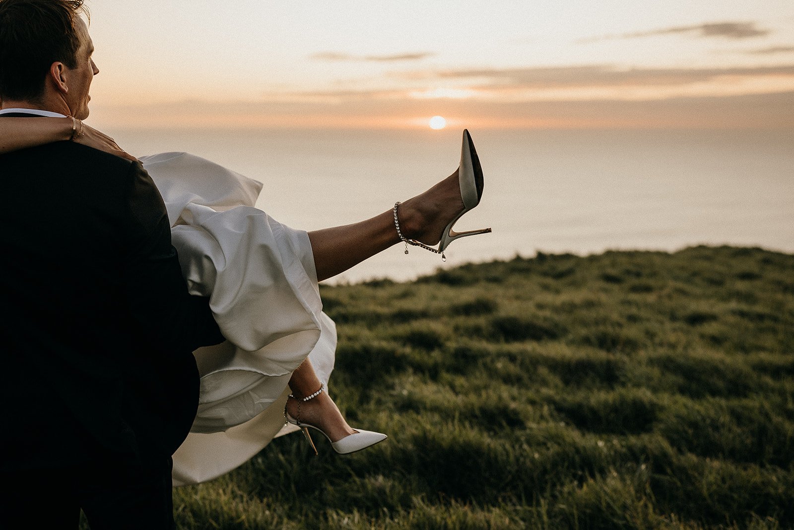 Man carrying a woman in a white dress across a grassy field at sunset