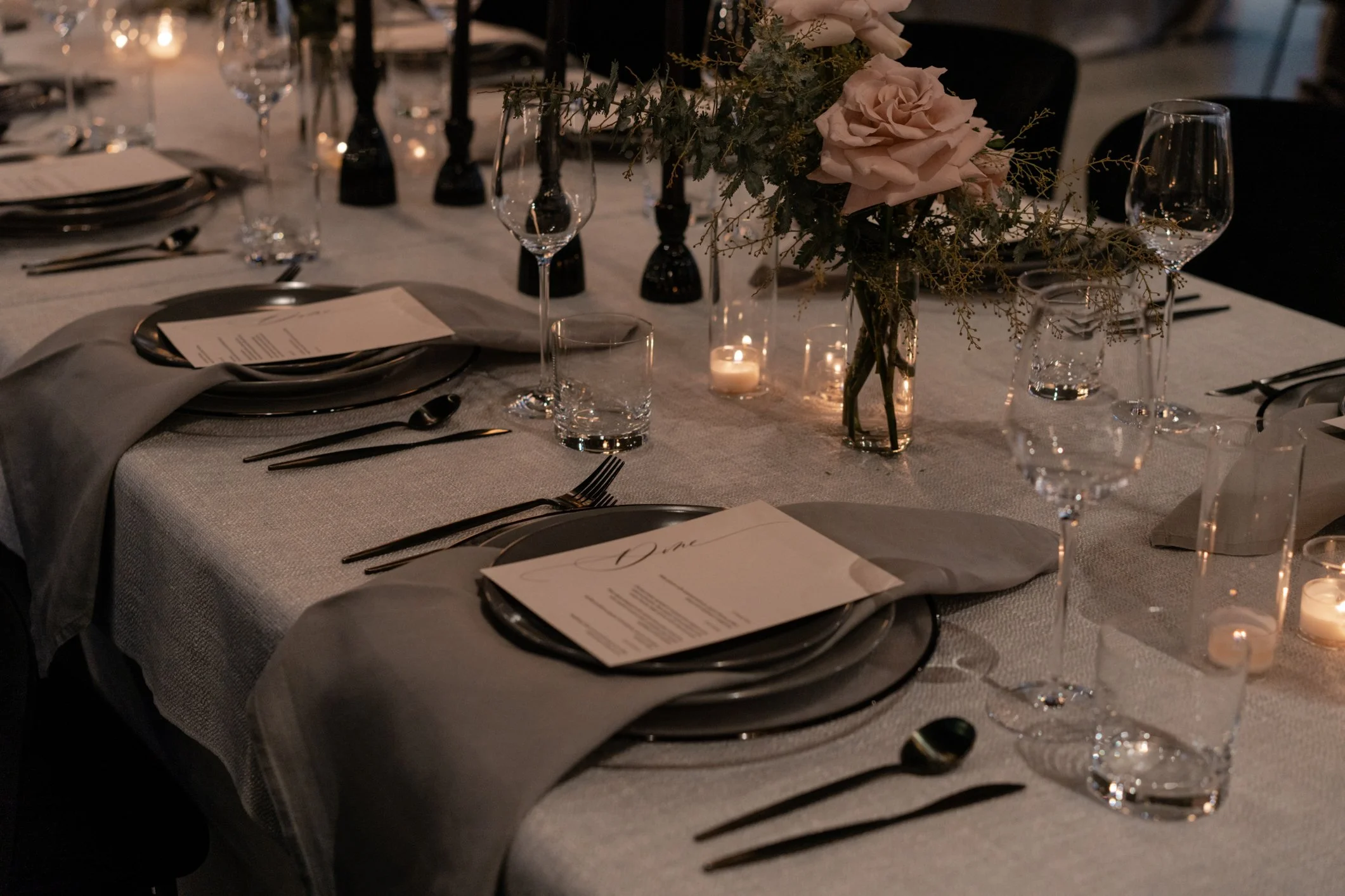 Elegant dinner table set with black plates, glassware, candles, menus, and a pink rose centerpiece