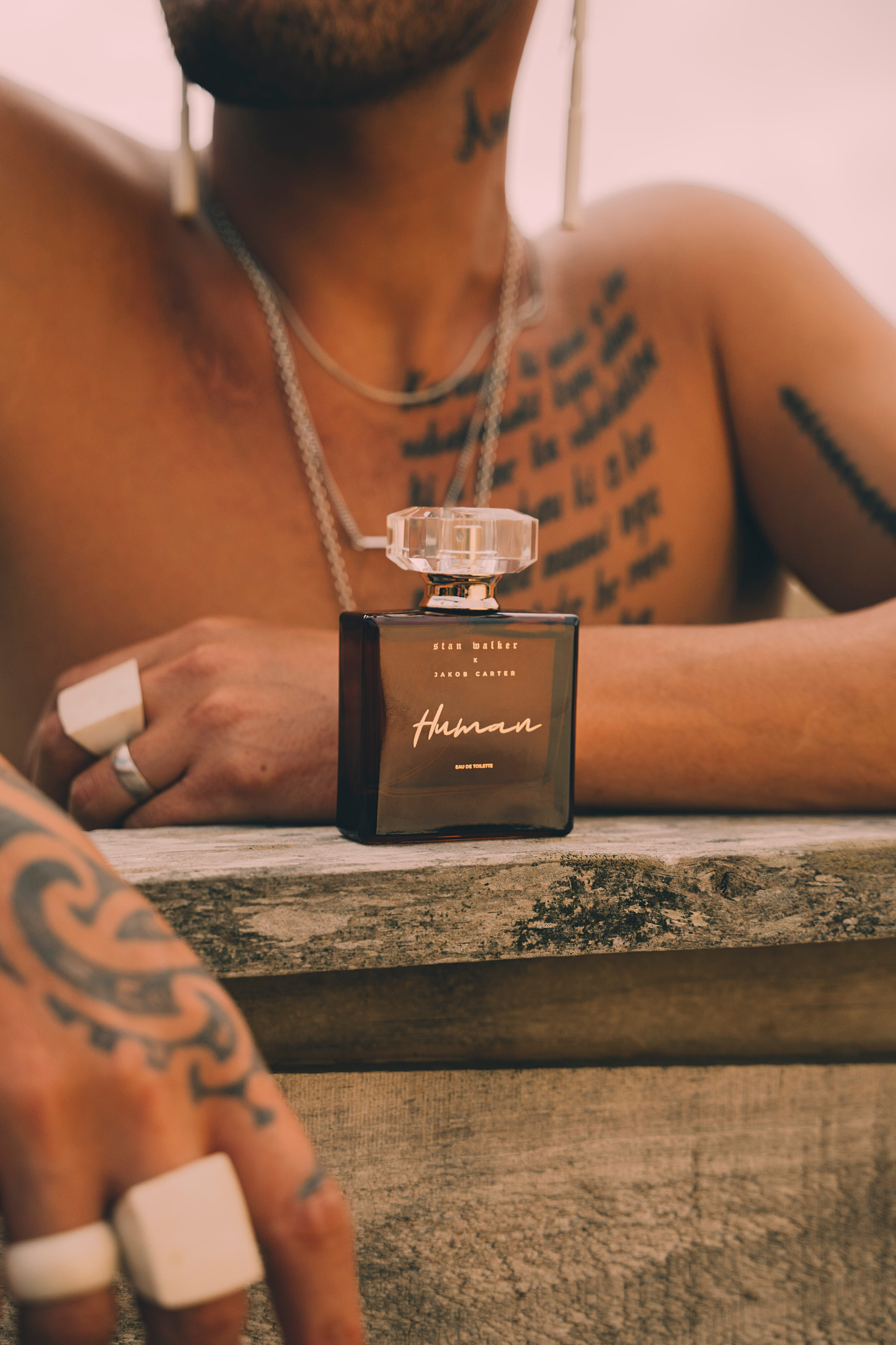 Stan Walker x Jakob Carter Human eau de toilette bottle on a wooden table in front of a tattooed person wearing layered necklaces