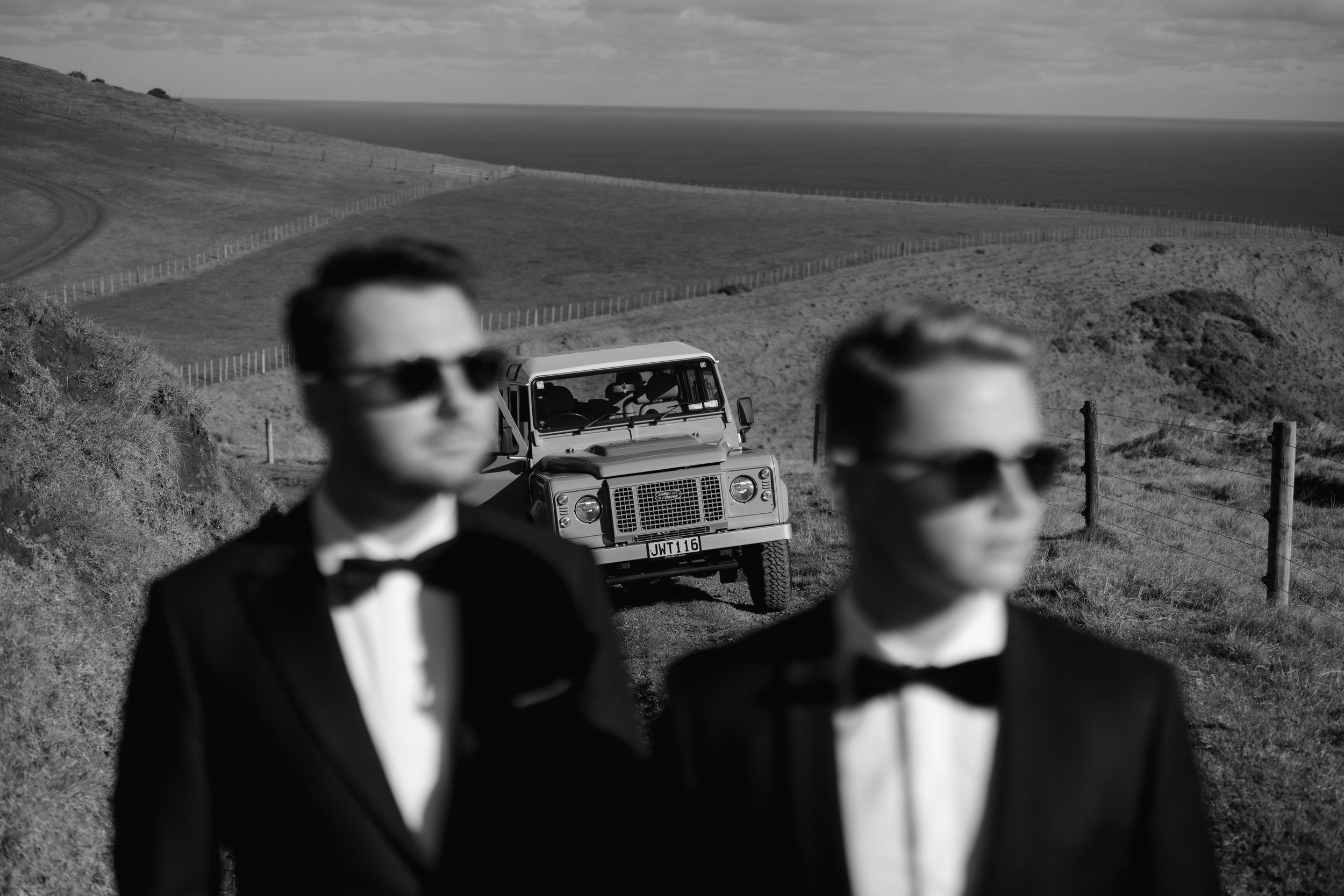 Black-and-white photo of two blurred men in tuxedos standing before a Land Rover on a fenced coastal hillside.