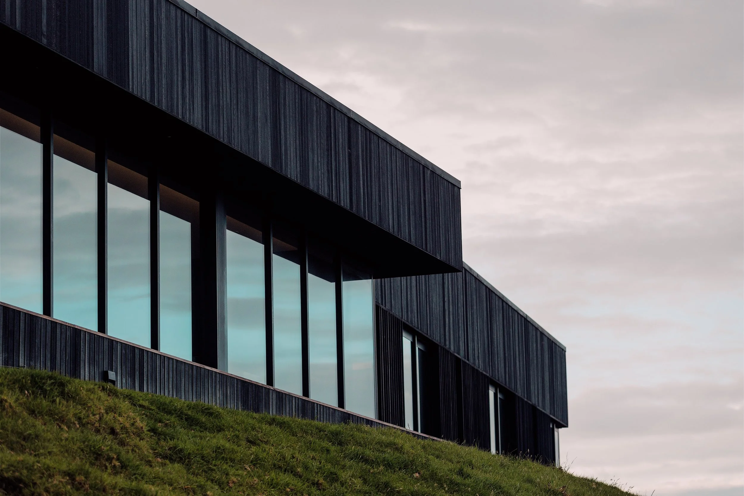 Modern dark building with large reflective windows on a grassy hillside beneath an overcast sky